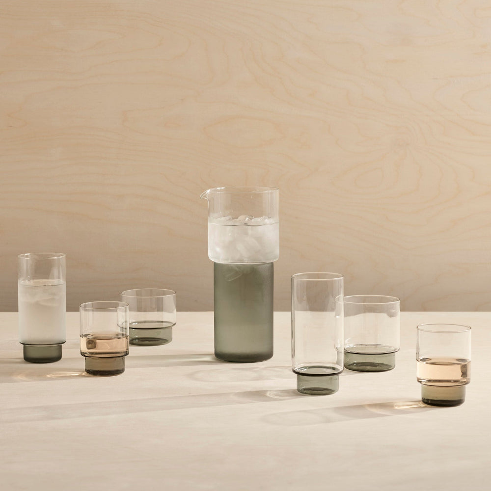 A set of smoked-tinted glass drinking glasses and a matching glass carafe filled with ice, arranged on a light wood tabletop against a light wood wall.