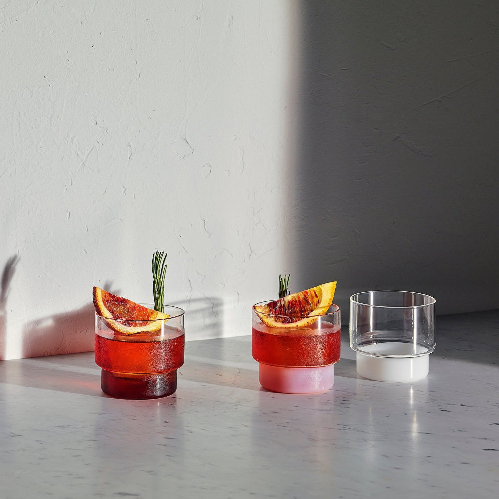 Three clear glass tumblers with stacked circular bases are arranged on a light stone surface against a white wall. Two tumblers are filled with a red drink and garnished with a blood orange slice and rosemary sprig, while the third tumbler is empty and clear.
