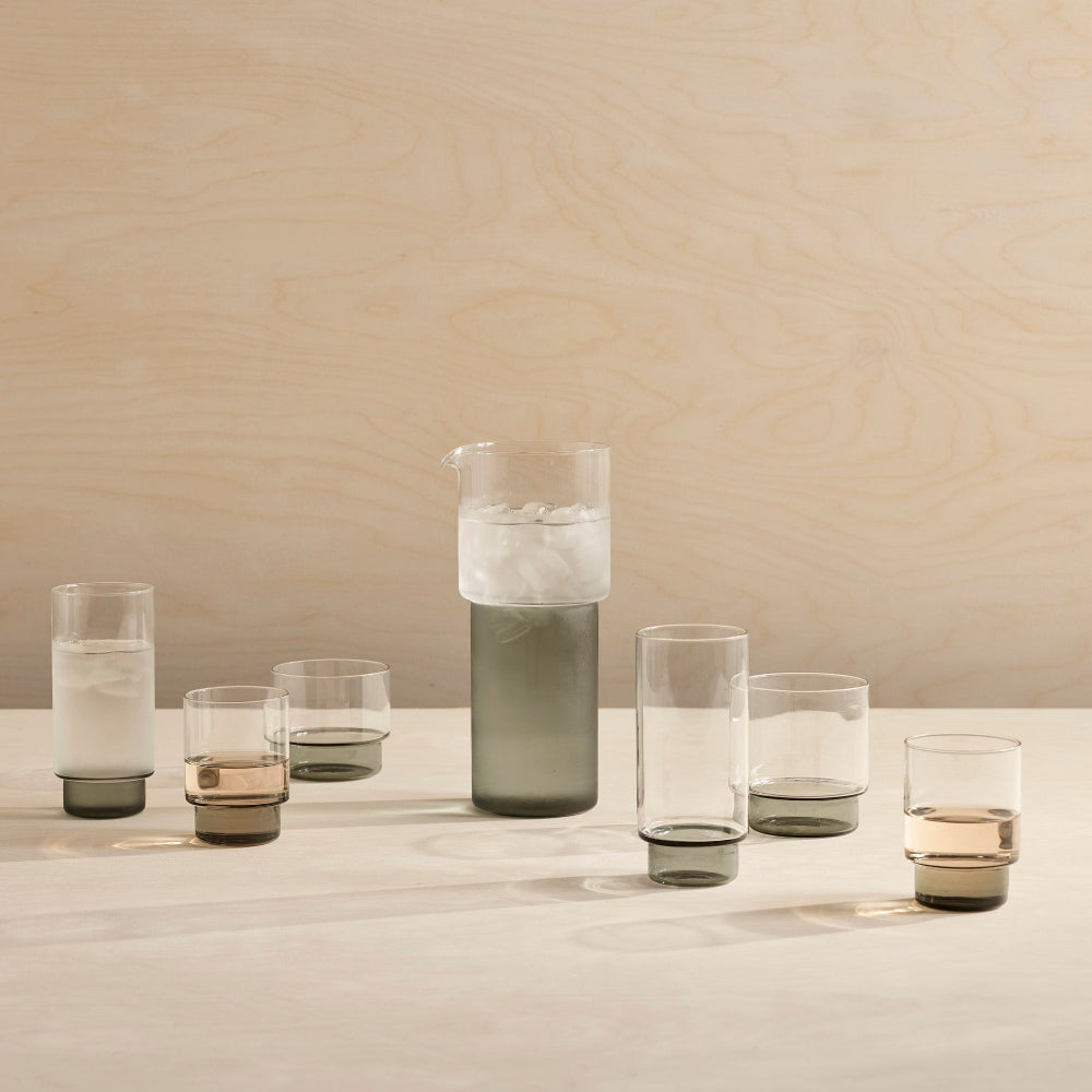A set of modern translucent drinking glasses and a matching carafe with smoky gray and clear glass sections, arranged on a light wood table against a beige wood-paneled background.