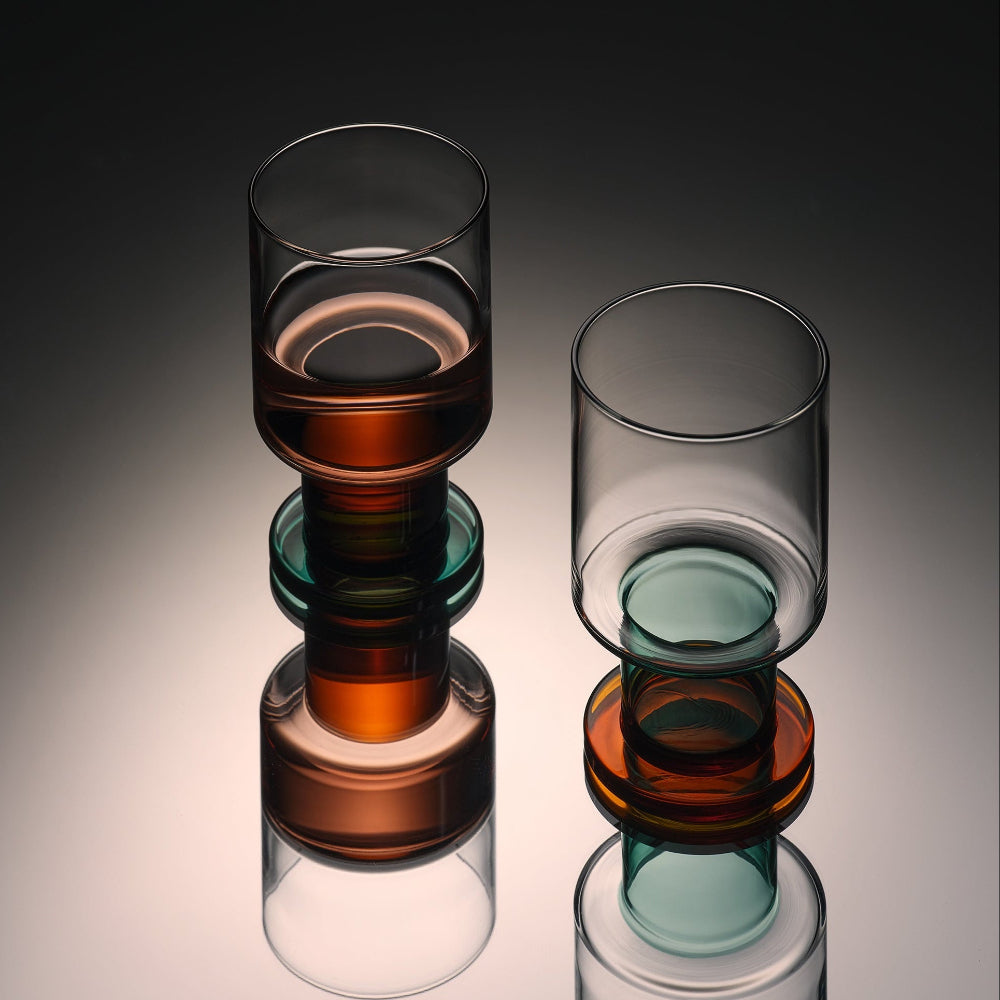 Two modern stacked glass tumblers with amber, teal, and smoky transparent layers, shown on a reflective dark gray studio background.