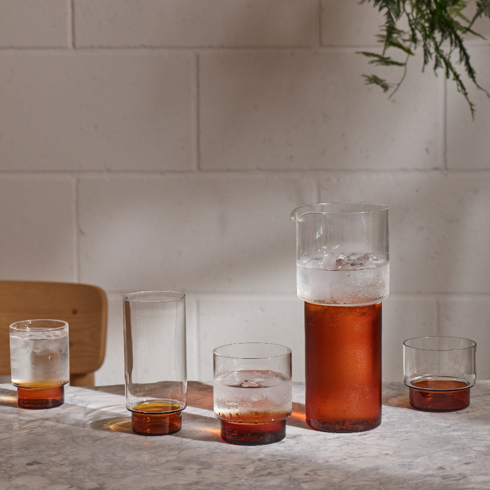 A set of clear glass drinking glasses and a matching pitcher with amber-tinted bases, arranged on a stone countertop against a light brick wall with natural sunlight and shadows.