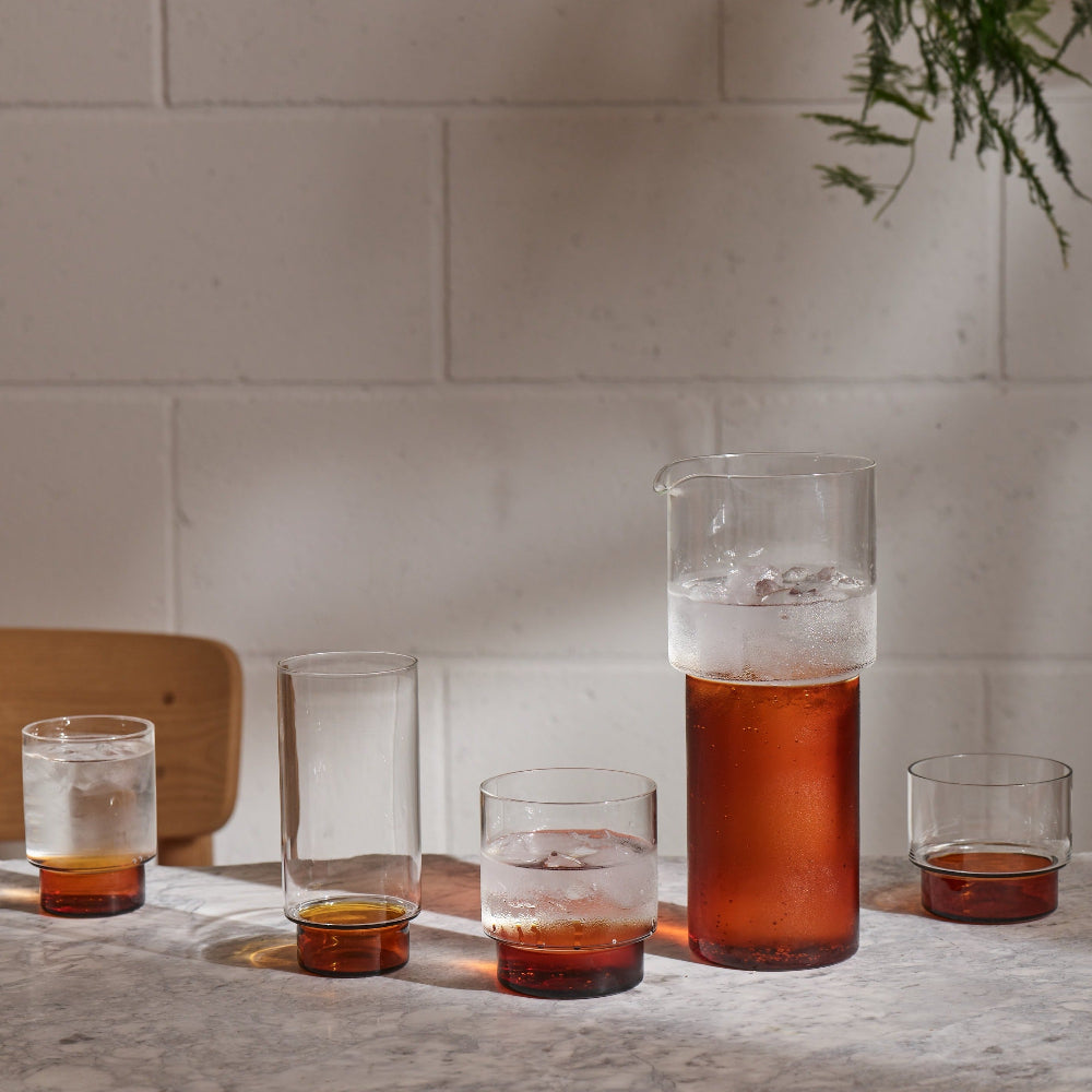 A set of clear glass drinking vessels including a tall pitcher and four tumblers, arranged on a stone surface against a light brick wall with natural sunlight and shadows.