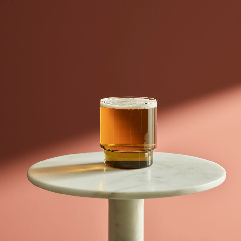 A clear lowball tumbler glass filled with amber beer and topped with white foam, set on a round white marble side table against a warm terracotta and peach background.