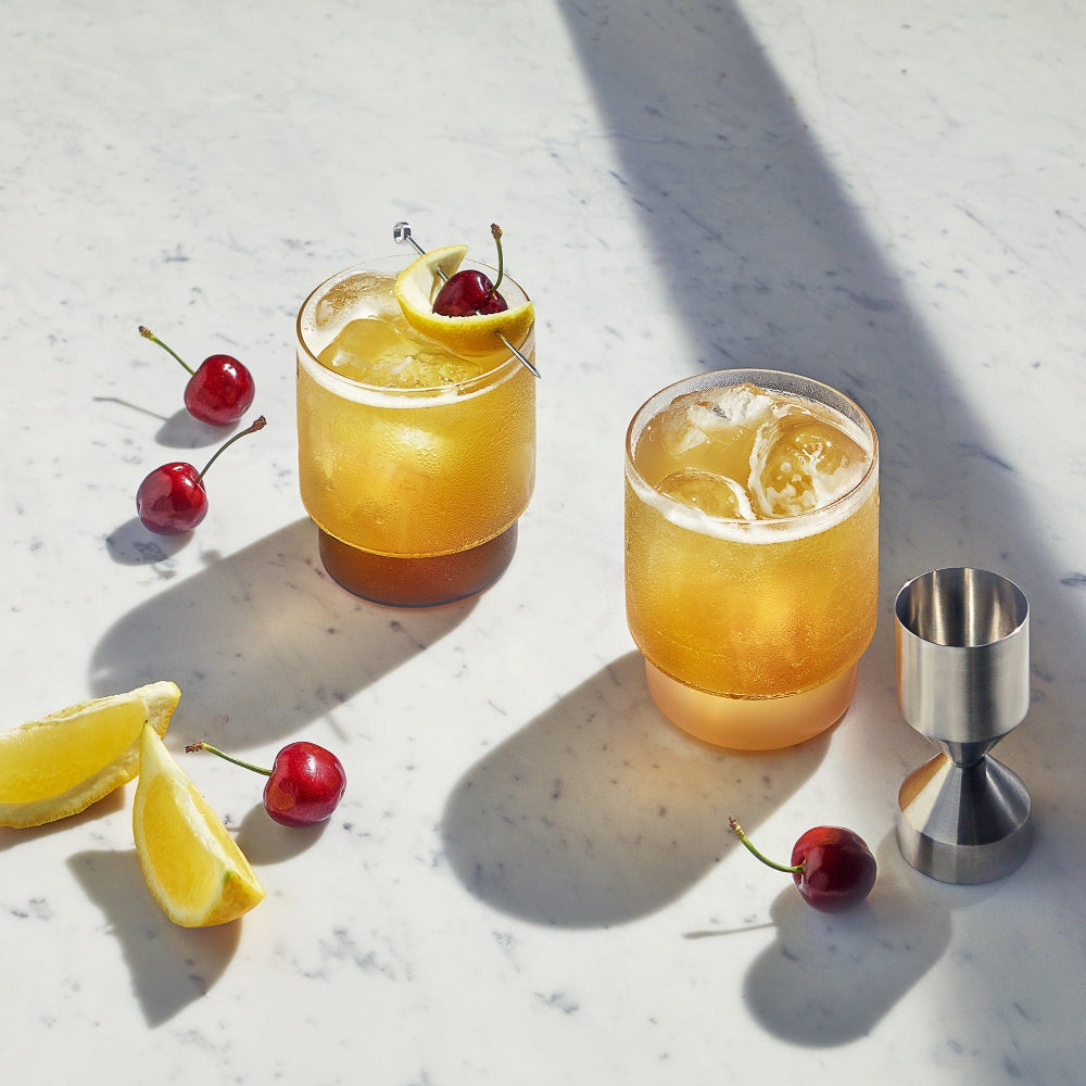 Two amber cocktails in clear rocks glasses with lemon wheels and cherries, arranged on a white marble surface with scattered cherries, lemon slices, and a stainless steel jigger under bright sunlight.
