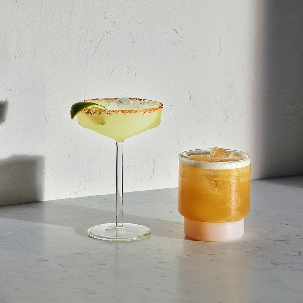 Two cocktails on a light marble surface against a textured white wall, with a yellow margarita in a coupe glass garnished with a lime wedge and an orange-colored iced drink in a short tumbler with a frosted top.