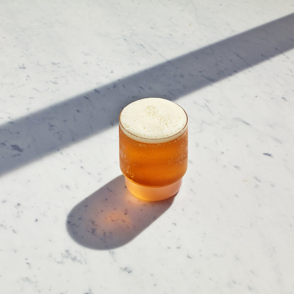 A frosted glass tumbler filled with an amber-orange beverage and topped with white foam, placed on a light stone surface in bright sunlight with a strong shadow.