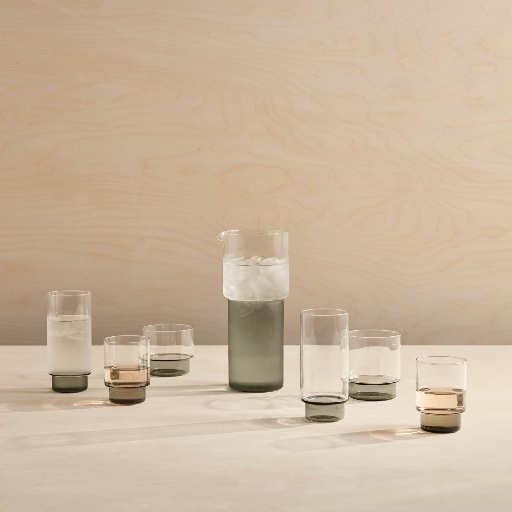 A set of six modern glassware pieces, including a tall carafe and five tumblers, arranged on a light wood surface against a matching wood-paneled background.