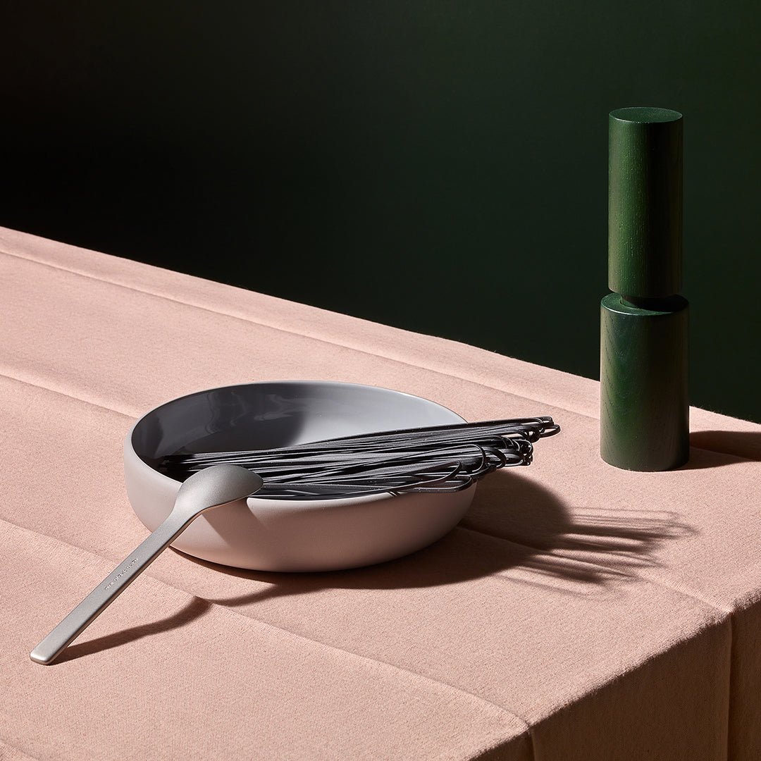 A matte white ceramic bowl with a gray interior, containing black chopsticks and a silver spoon, set on a dusty pink tablecloth next to a dark green pepper mill against a dark green background.