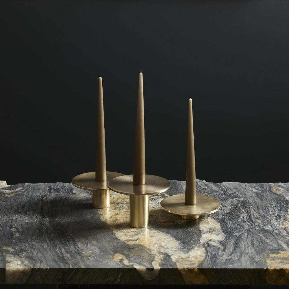 Three brass taper candle holders with conical candles, displayed on a dark marble surface against a solid black background.