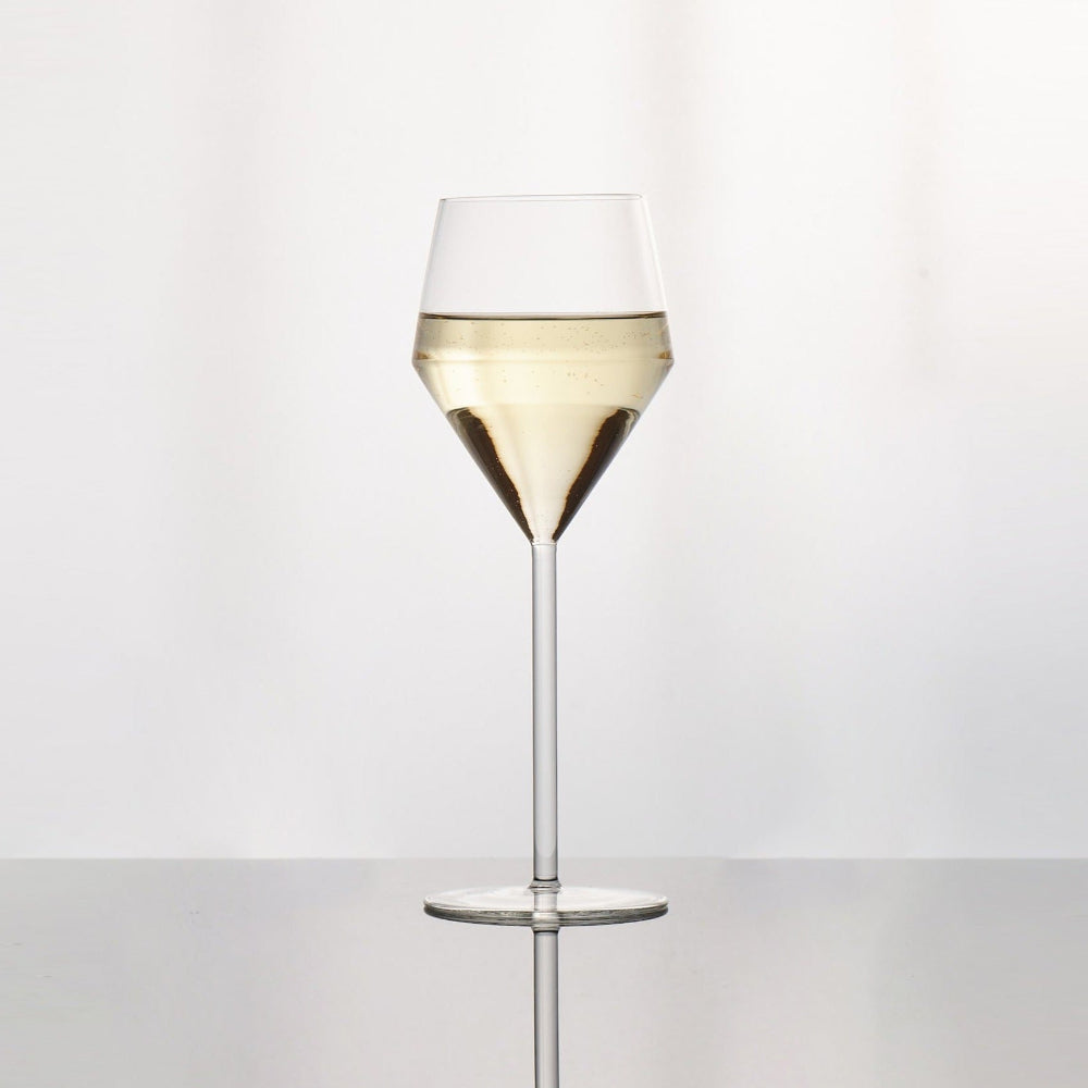 A clear glass wine glass with a wide bowl, filled with white wine, standing on a reflective surface against a light gray background.