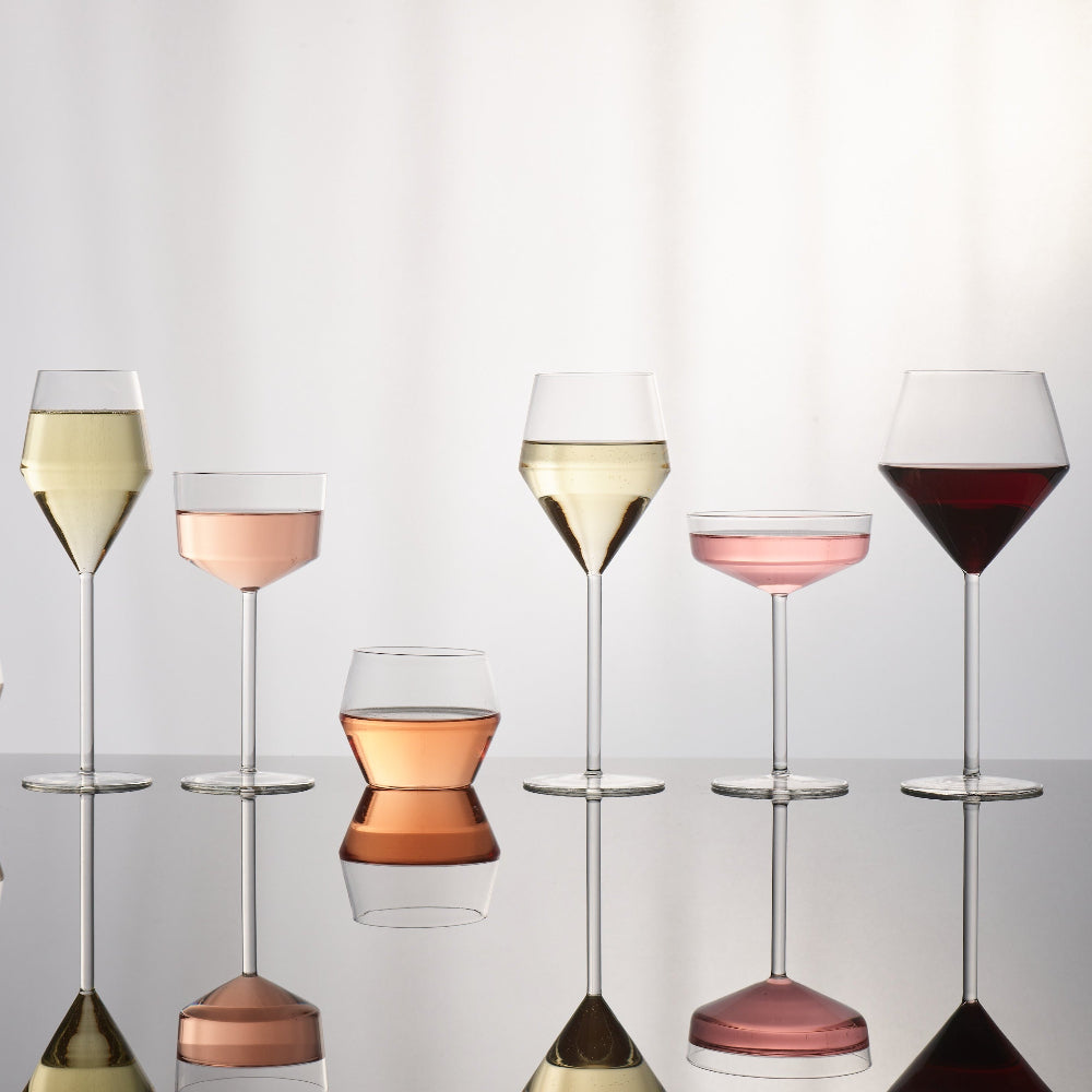 Six clear wine and cocktail glasses in different shapes, some filled with white wine, pink wine, and red wine, arranged on a reflective surface against a soft light gray background.
