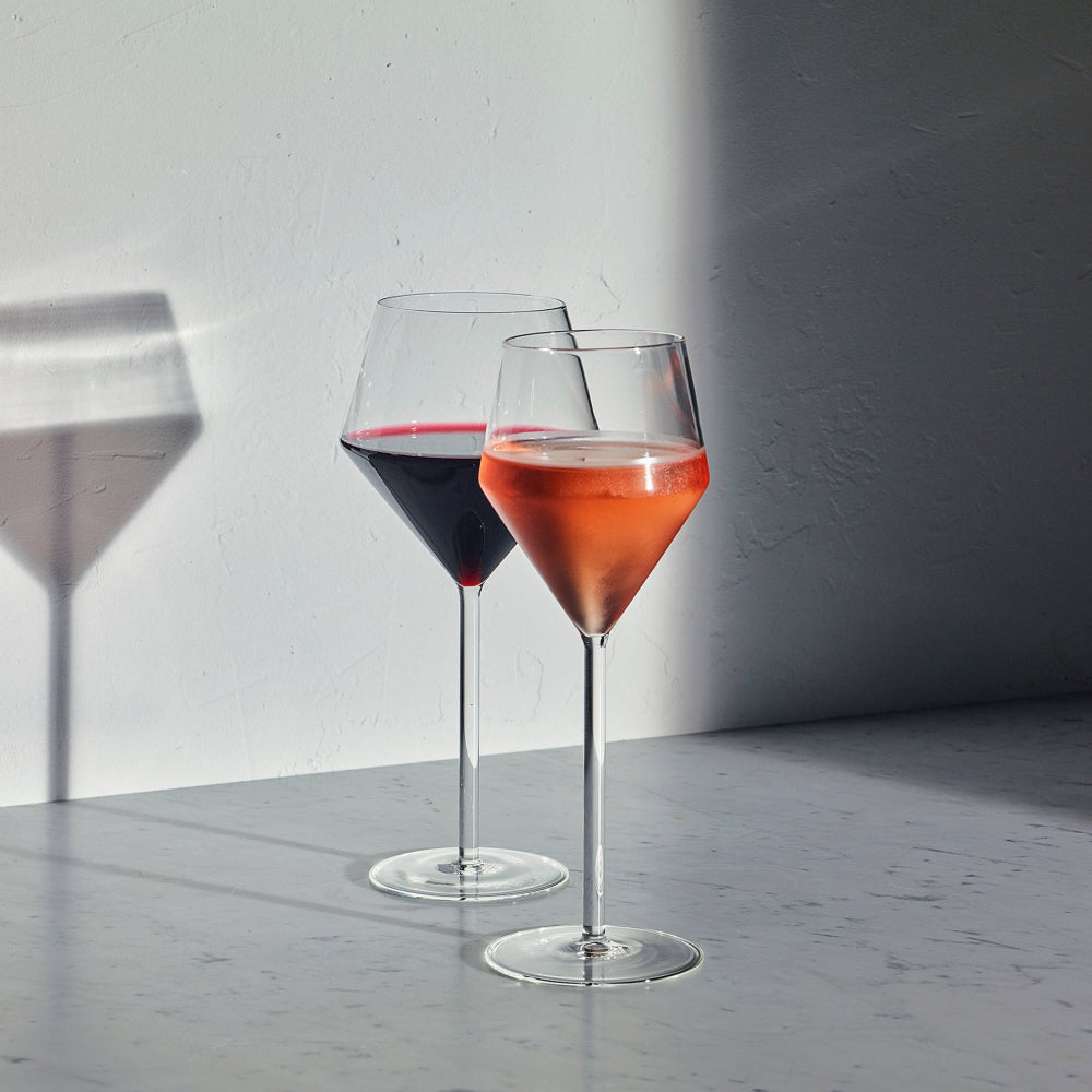 Two clear stemmed wine glasses on a gray surface against a white wall, one filled with red wine and the other with a pink drink, with strong sunlight casting shadows.