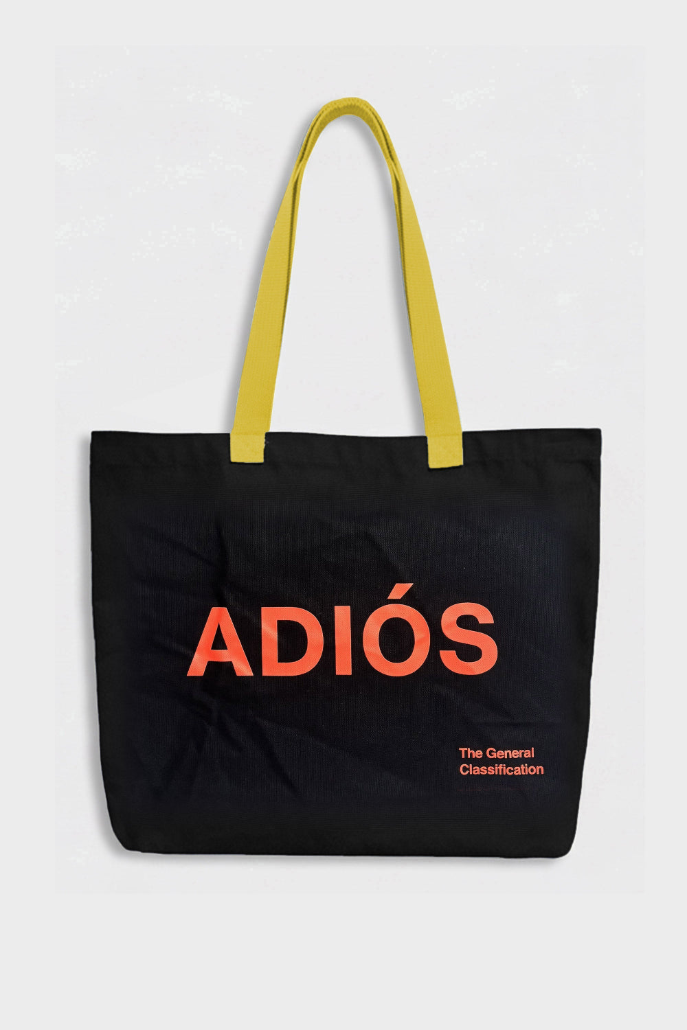 A black canvas tote bag with bright yellow shoulder straps and bold orange-red text reading 'ADIÓS' on the front, plus smaller text 'The General Classification' near the lower right. The bag is photographed against a light grey background.