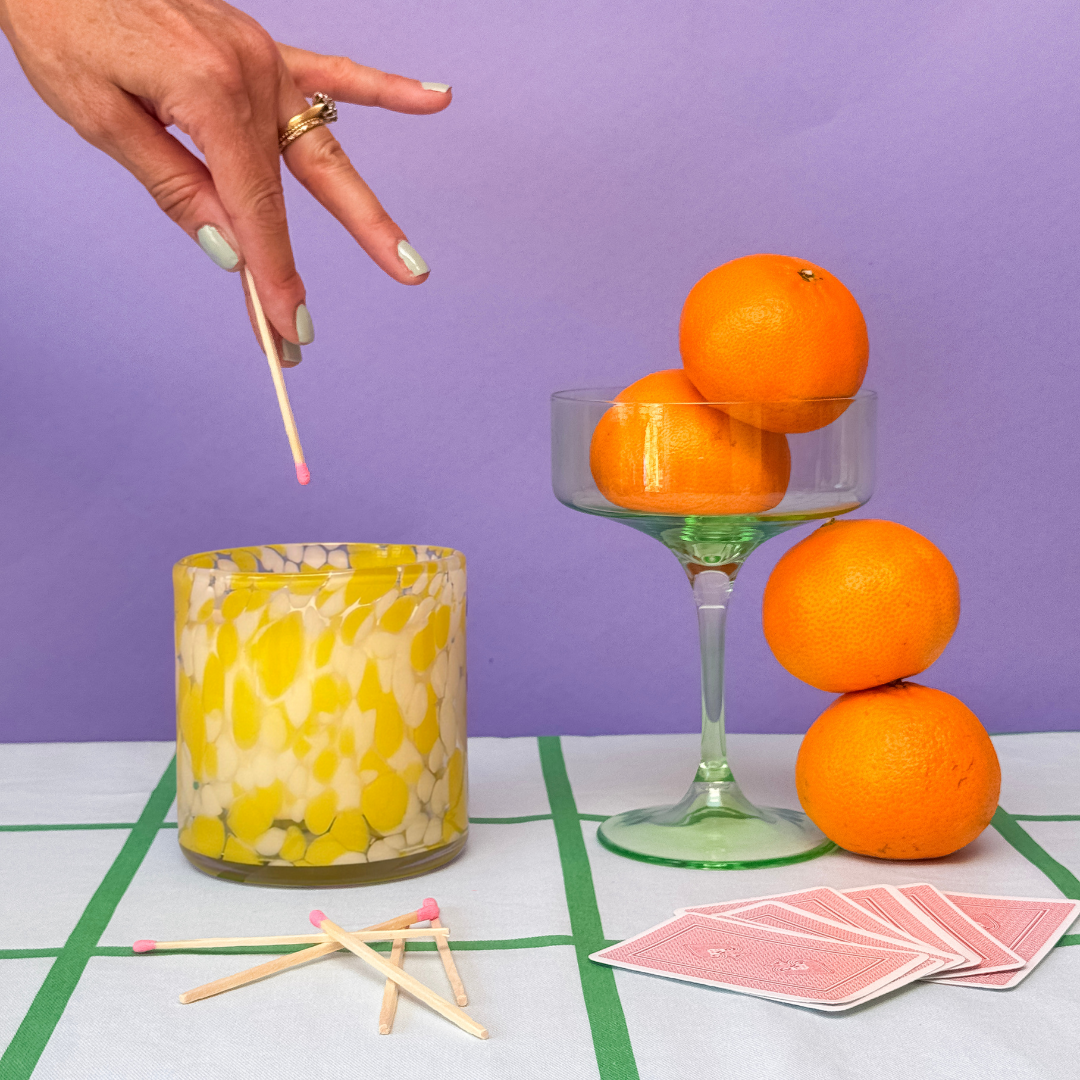 Yellow candle with lemon design, oranges, and playing cards on a checkered tablecloth against a purple background.