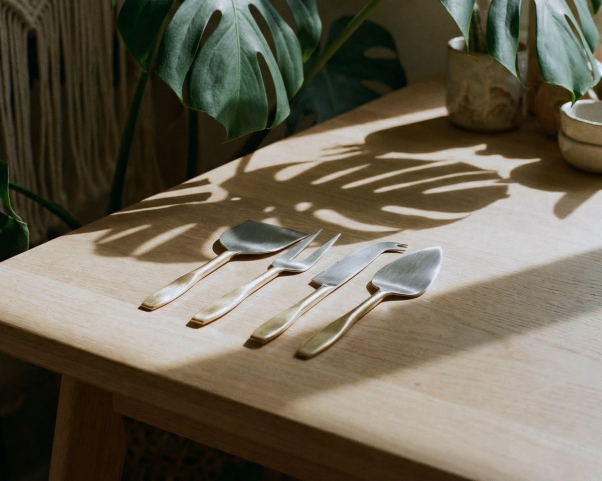 Set of four silver cheese knives on a wooden table with plant shadows