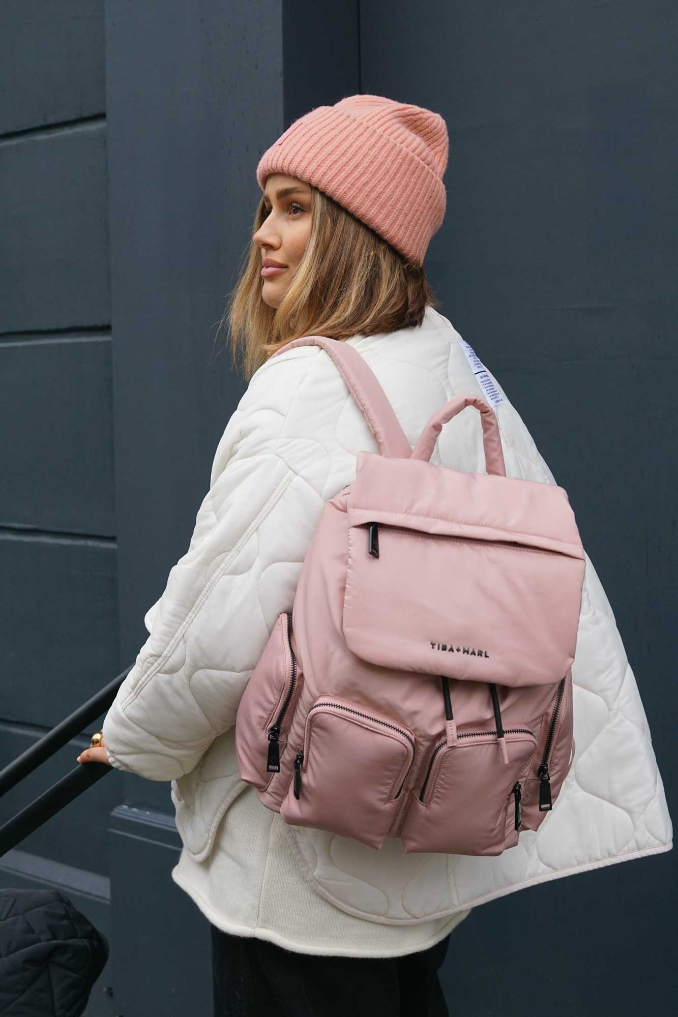 Woman wearing a pink beanie and carrying a pink backpack against a dark background pushing a baby buggy.