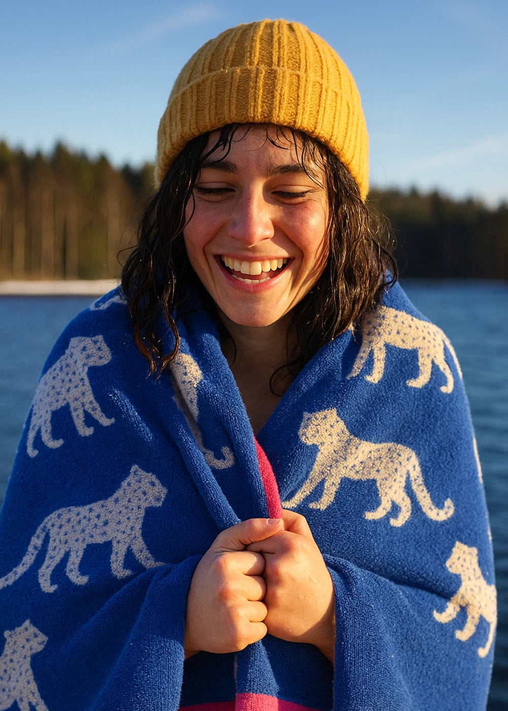 Person wrapped in a blue blanket with animal patterns, wearing a yellow beanie, standing by a body of water.