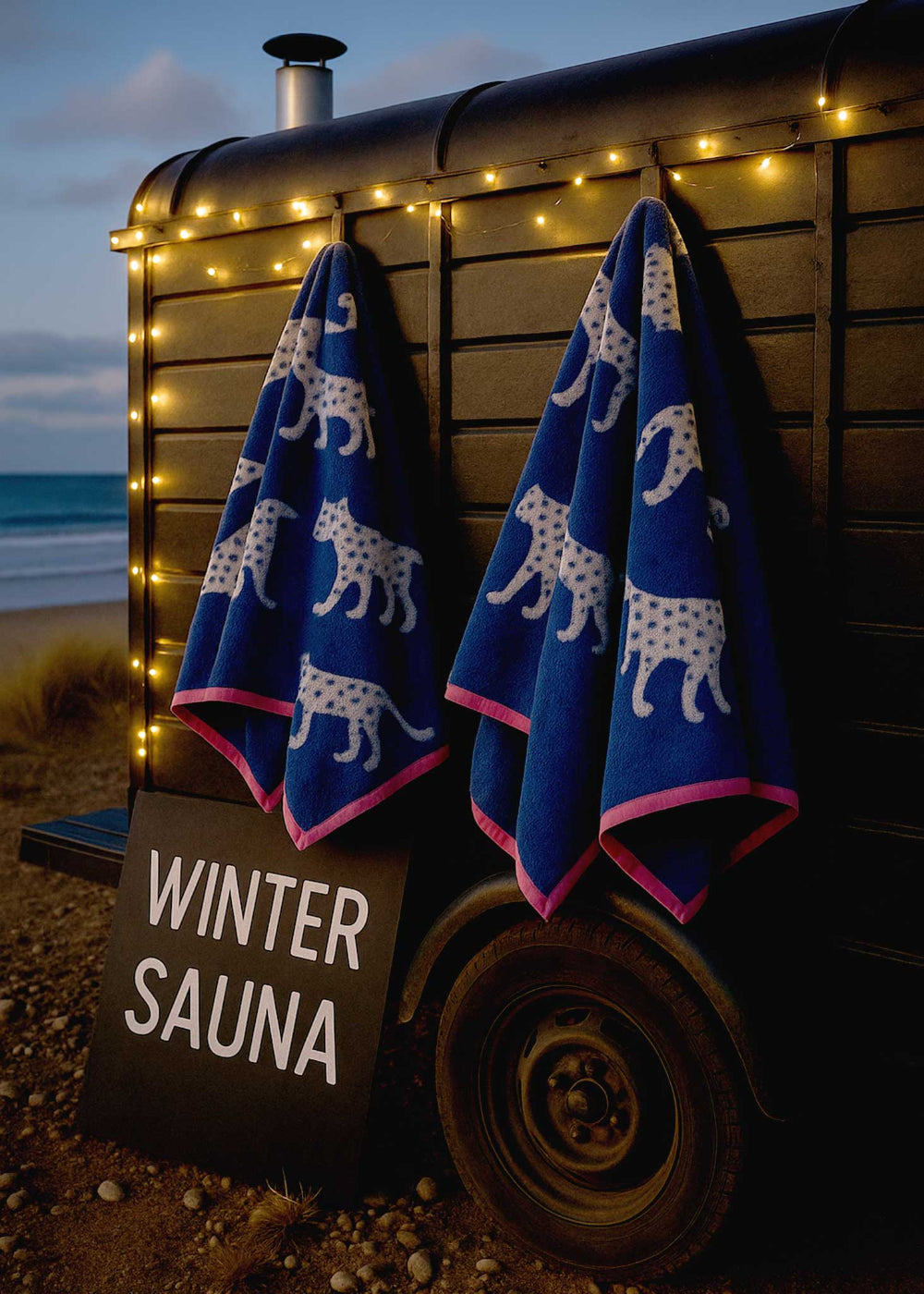 Two blue towels with white animal patterns hanging on a wooden structure by the beach at dusk.