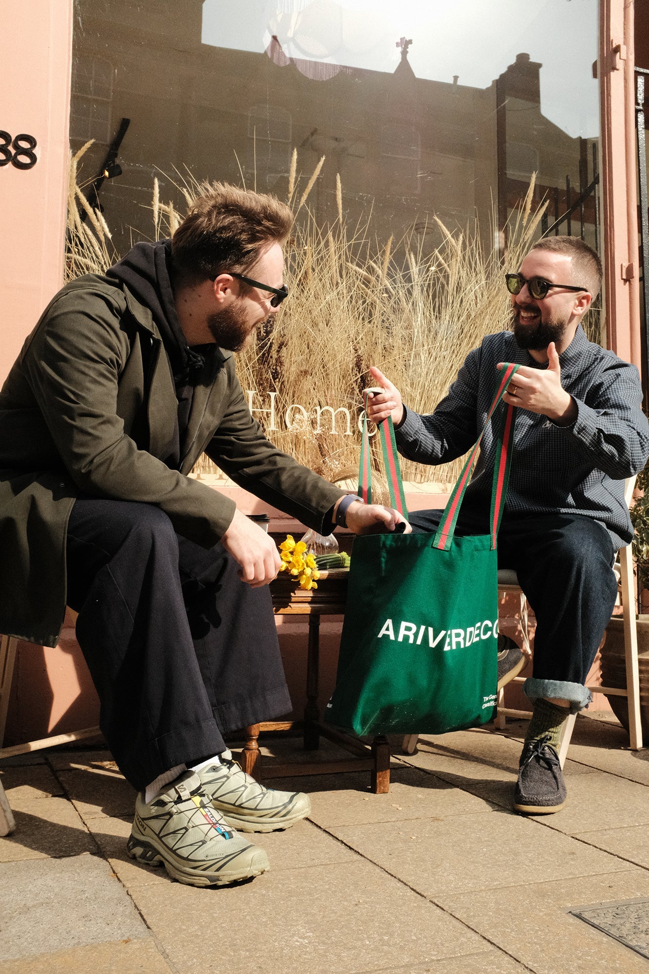 Two men sitting outdoors, one handing a green bag to the other with 'ariverderci' branding.