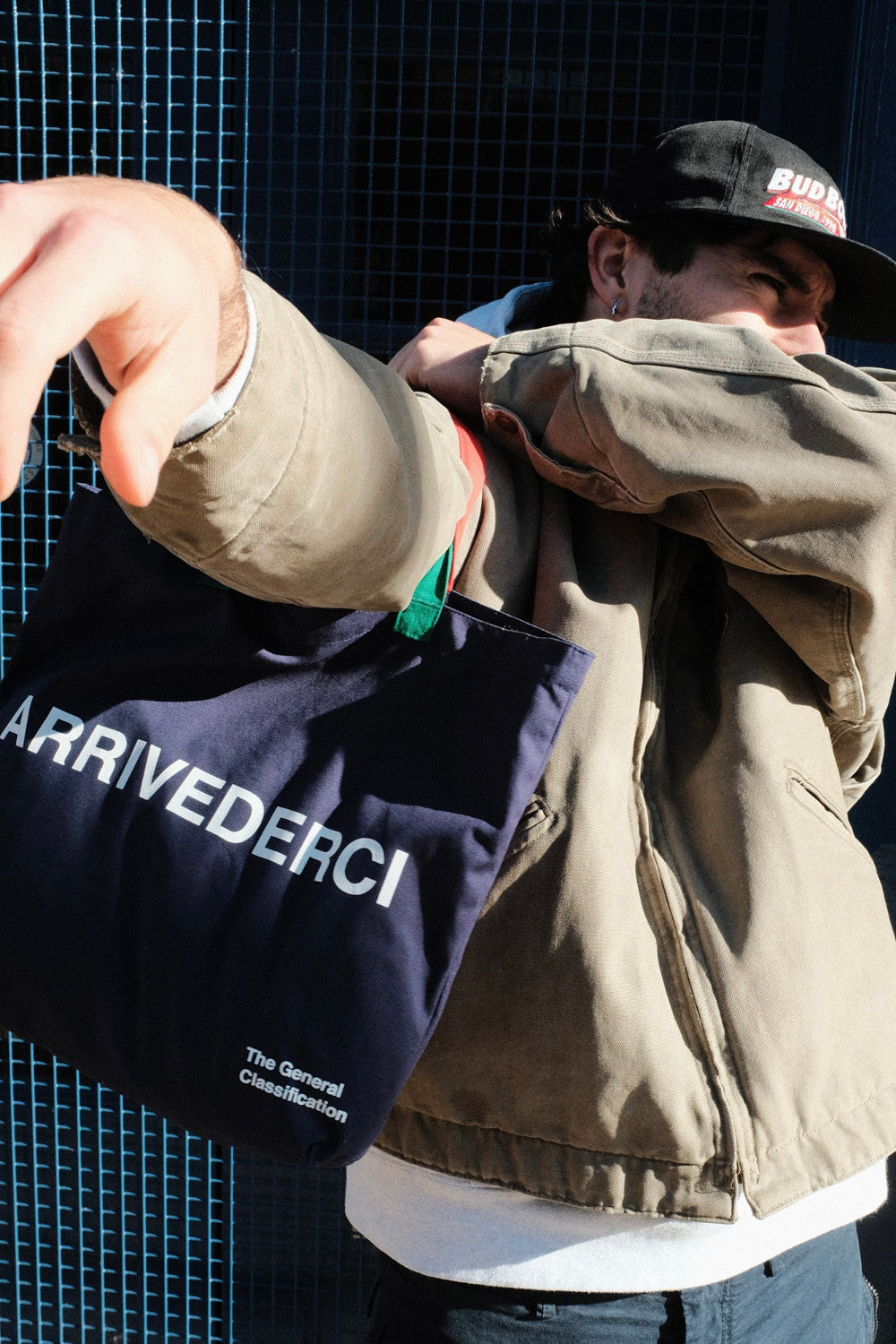 A person wearing a beige jacket and black cap holds a navy blue tote bag with the brand name 'ARRIVEDERCI' printed in white. The background features a dark blue grid-patterned wall.