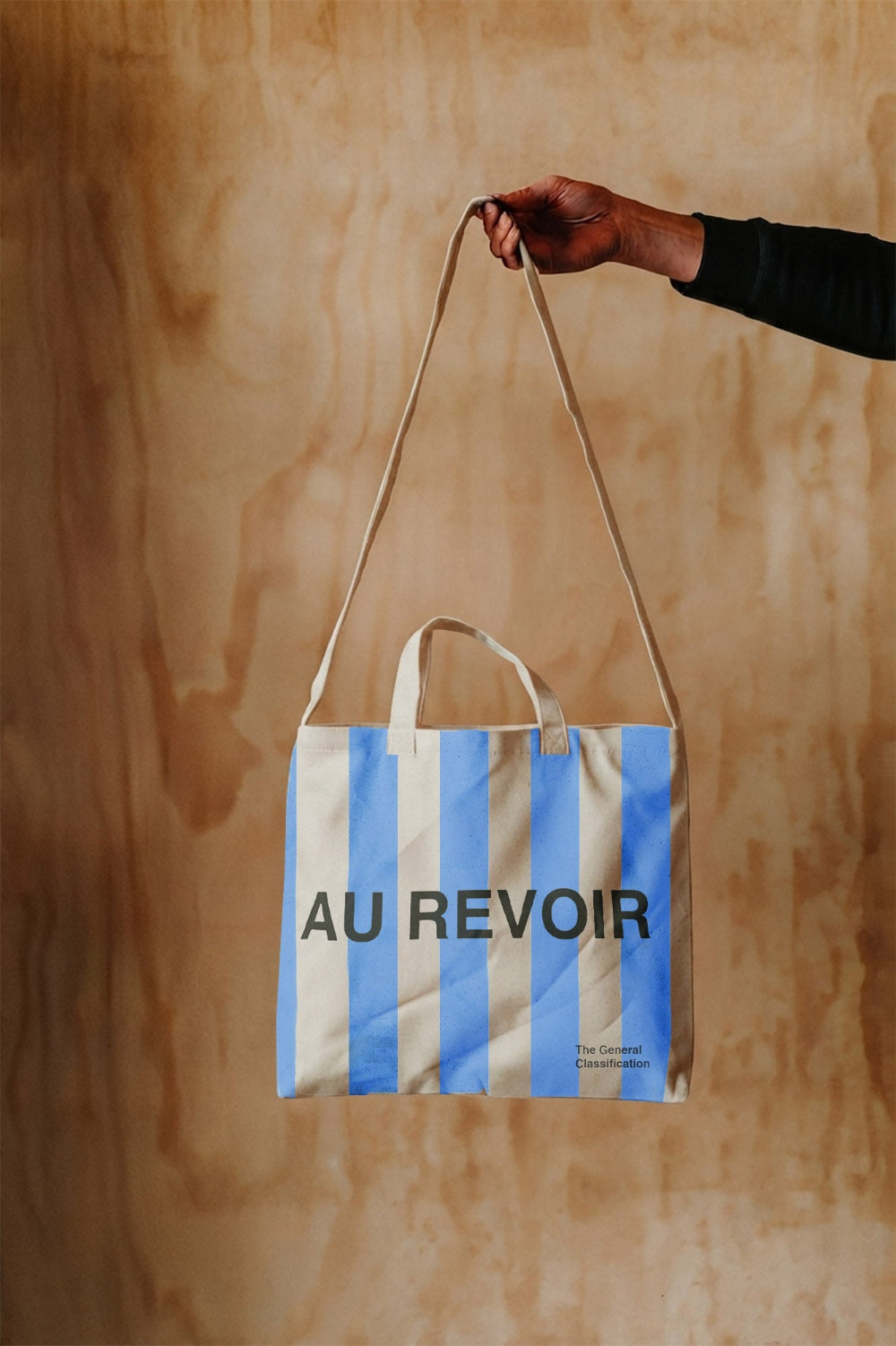 A hand holding a canvas tote bag with blue and white vertical stripes and the text 'AU REVOIR' printed in black, set against a light wood background.
