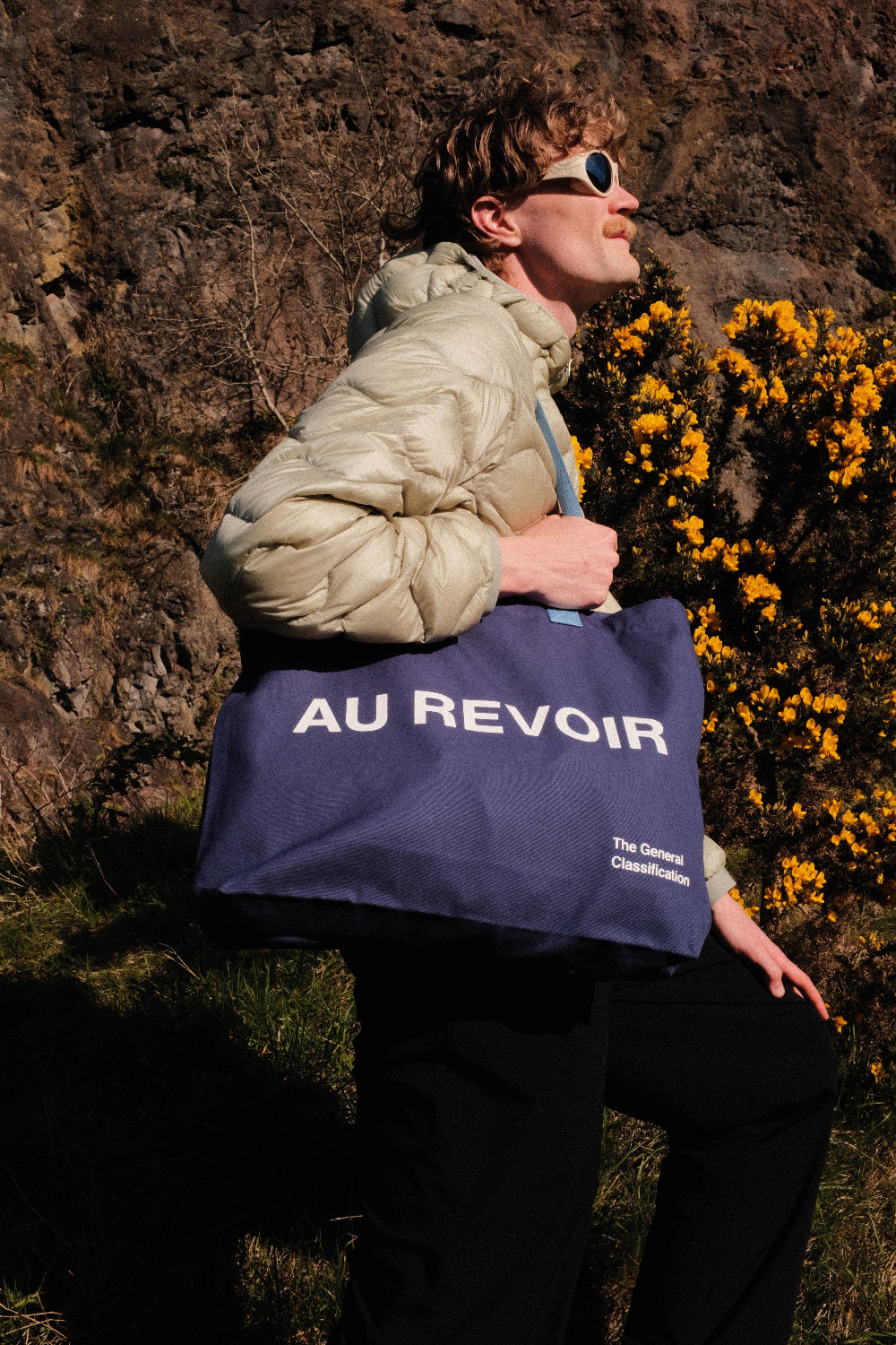 Person holding a blue tote bag with 'AU REVOIR' text against a natural background