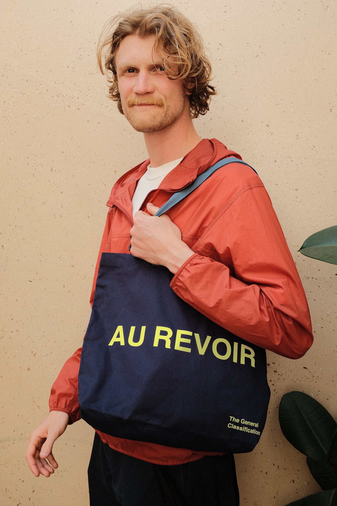 Man wearing a red jacket with a blue bag labeled 'AU REVOIR' against a beige wall.
