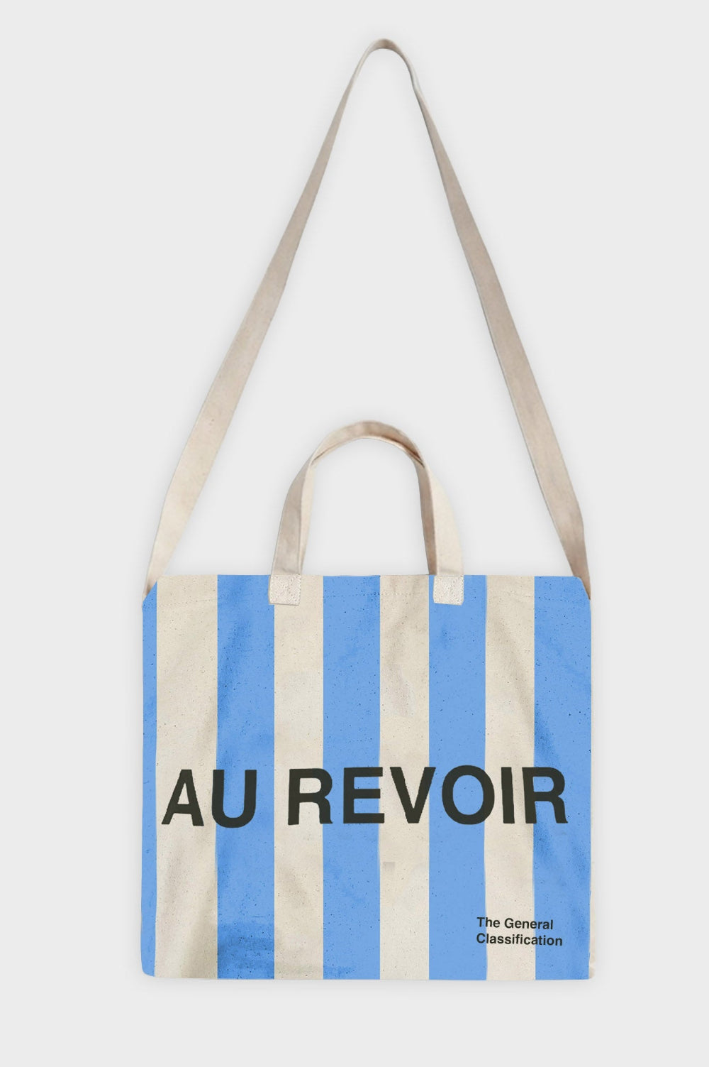 A canvas tote bag with vertical light blue and cream stripes, featuring the text 'AU REVOIR' in black across the center and 'The General Classification' in the bottom right corner. The bag has beige fabric handles and is photographed against a light grey background.