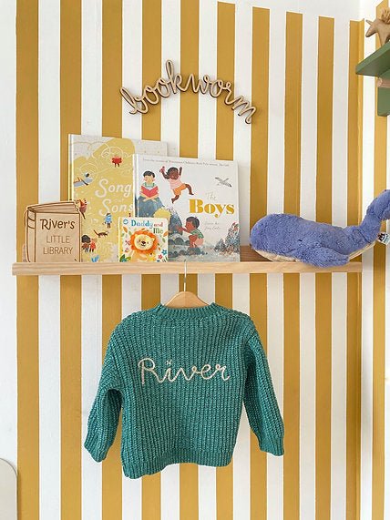 Children's sweater with 'River' embroidery, books, and a toy on a shelf against a striped wall.