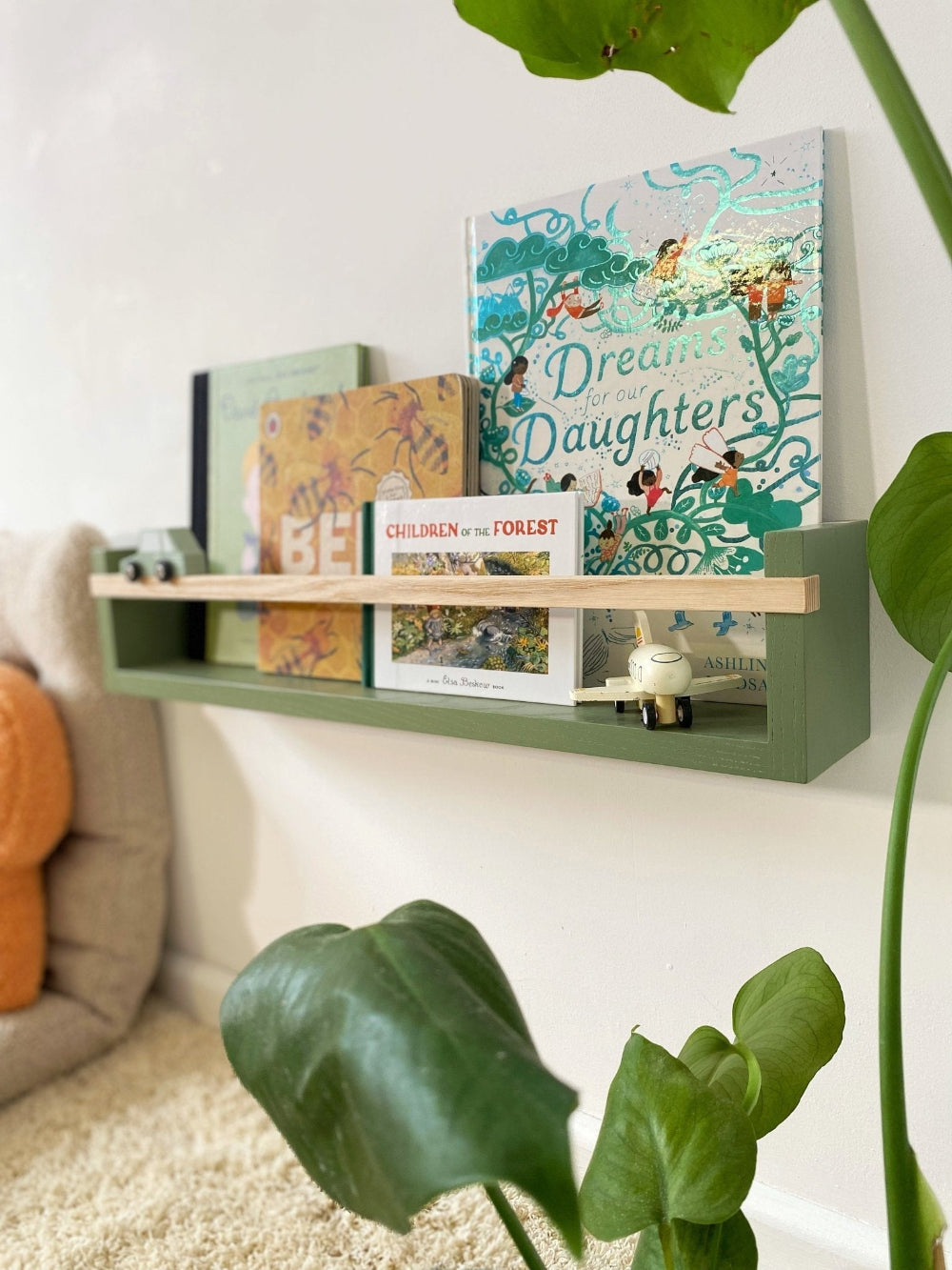 Green shelf with books and decorative items against a white wall, surrounded by plants.