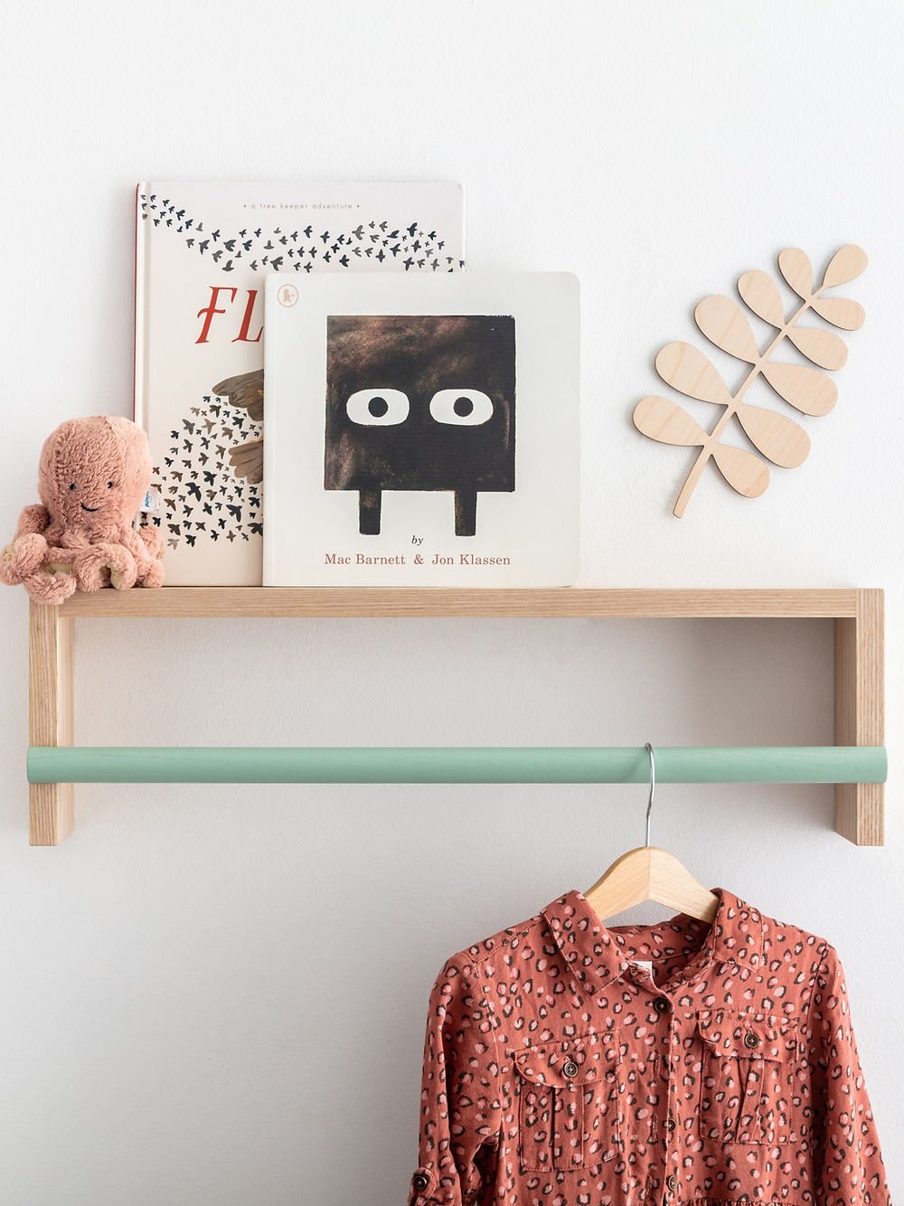 Wooden shelf with books, a teddy bear, and a floral shirt on a hanger against a white wall.
