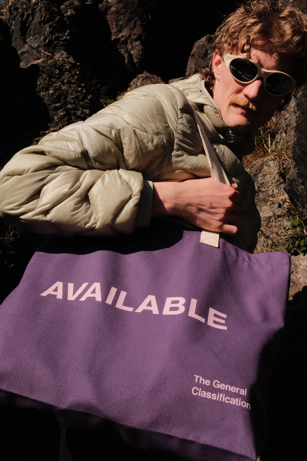 Person holding a purple tote bag with 'AVAILABLE' text against a dark background