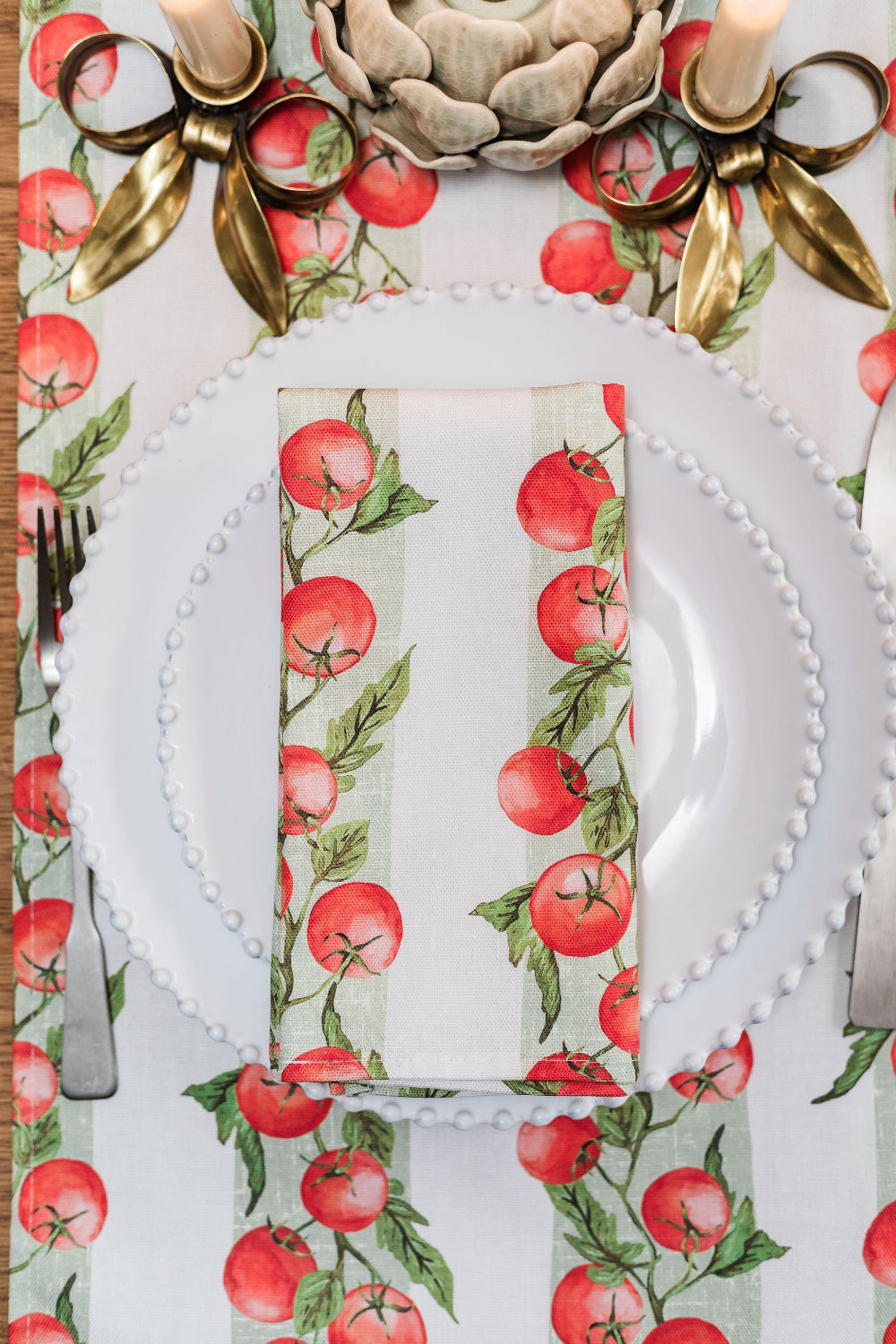 Table setting with white plates, napkins, and cutlery on a decorative tablecloth with tomato vine pattern.