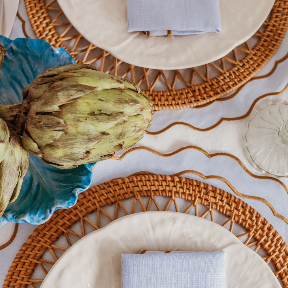 Artichoke on a blue plate with woven placemats and white plates on a table.
