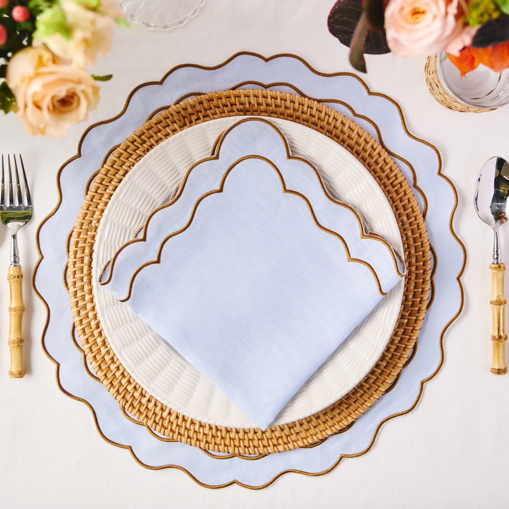 Decorative woven placemat with baby blue scalloped edge placemat on a white tablecloth, surrounded by floral arrangements and cutlery.