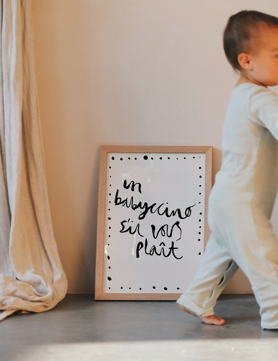Child standing next to a framed poster with 'un babyccino' text on a neutral background