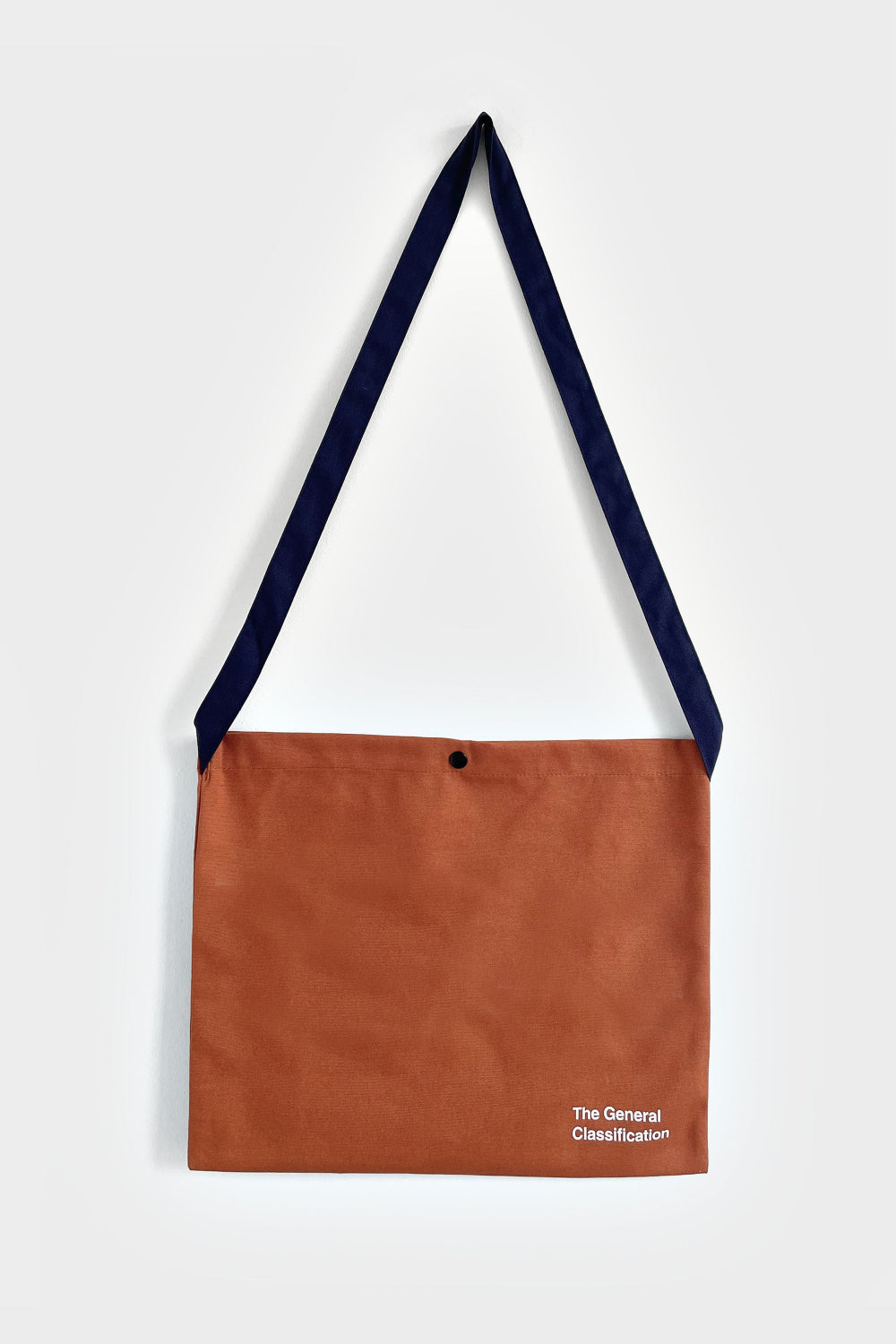 A brown shoulder bag with a navy blue strap and white text reading 'The General Classification' on the bottom right corner, shown against a light grey background.
