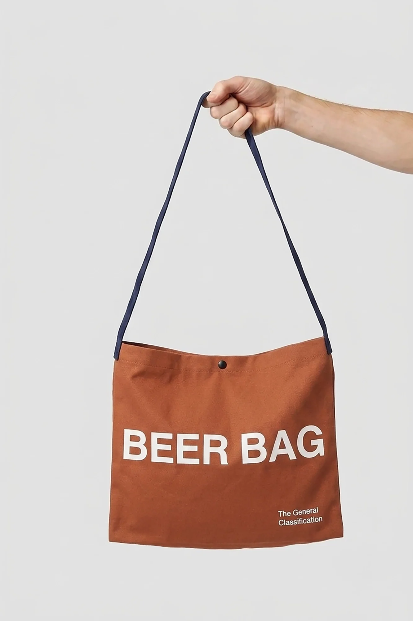 A brown canvas shoulder tote bag with a navy blue strap, held by a person's hand against a light gray background. The bag features the text 'BEER BAG' in white capital letters and 'The General Classification' in smaller white text near the bottom right.