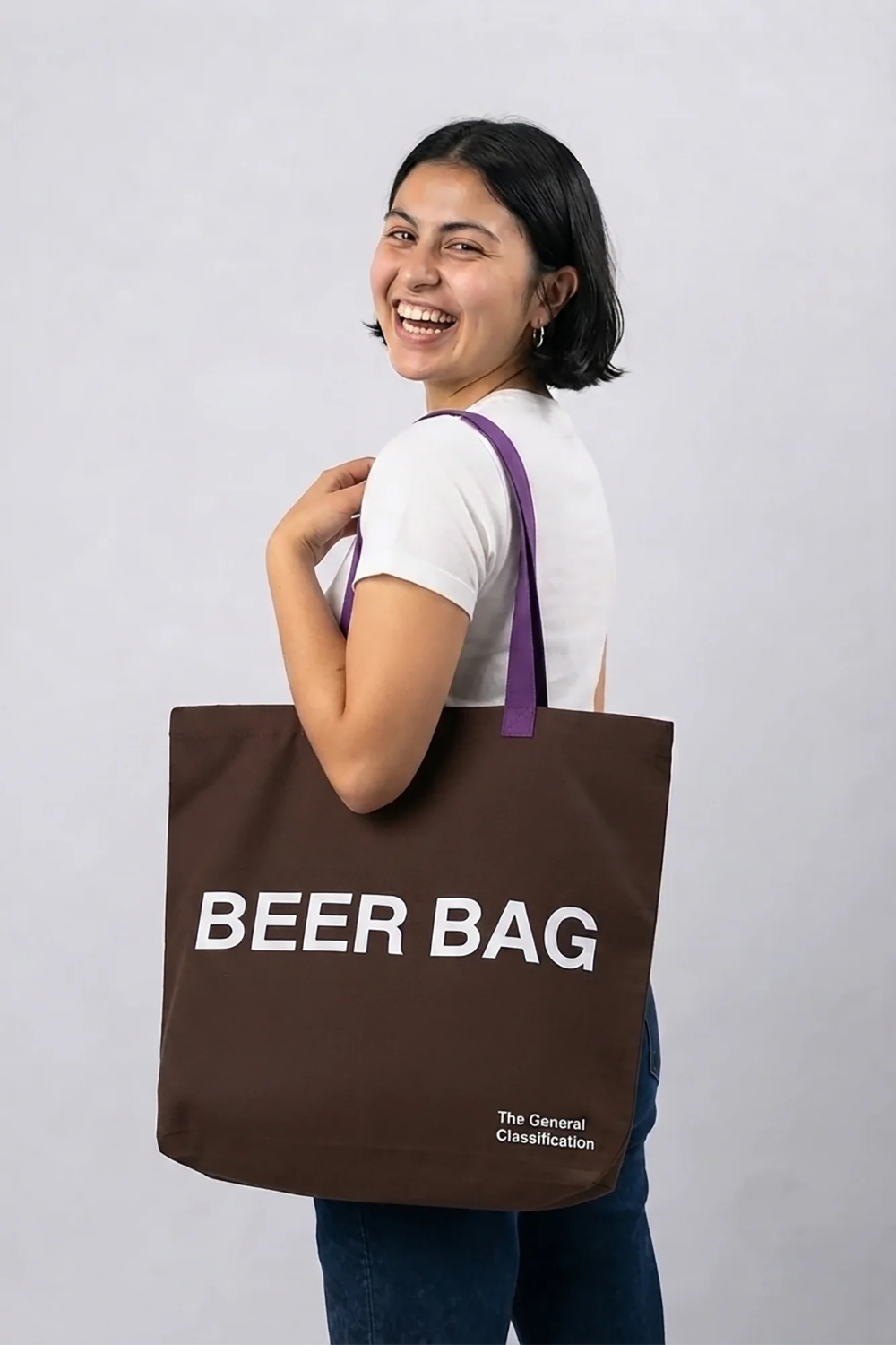 A woman smiling and looking back over her shoulder while carrying a brown canvas tote bag with purple handles. The bag features the text 'BEER BAG' in bold white letters and 'The General Classification' in smaller white text near the bottom right.