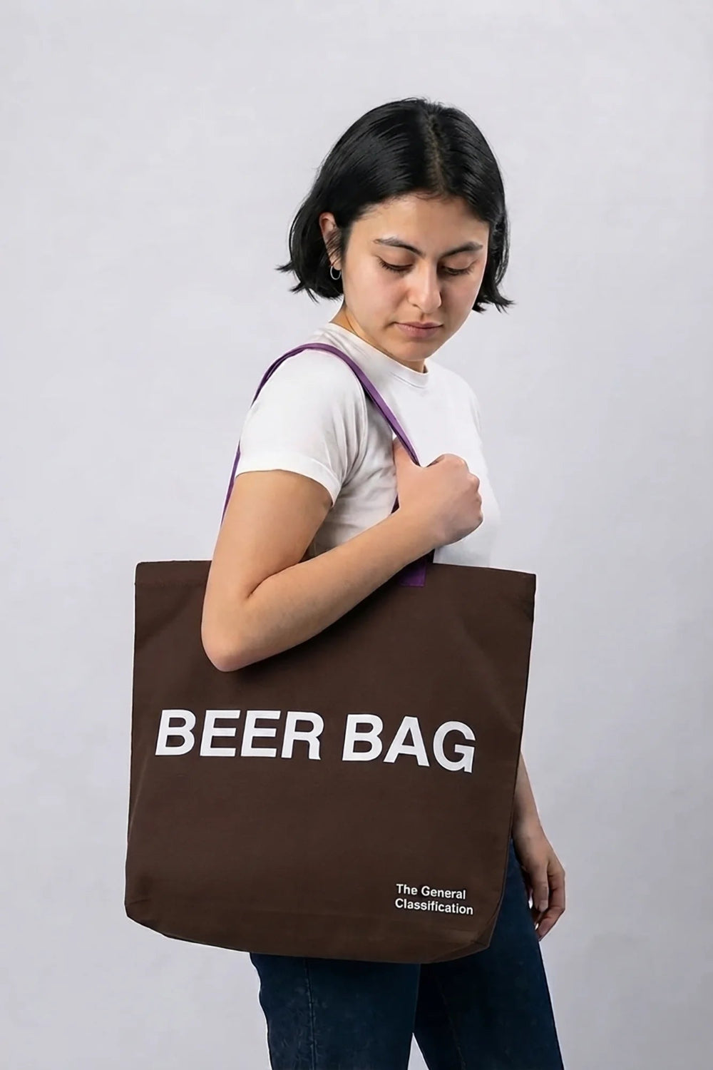 A woman wearing a white t-shirt and blue jeans carries a brown canvas tote bag with purple shoulder straps. The bag features the text 'BEER BAG' in bold white letters, with 'The General Classification' printed in small white text below.