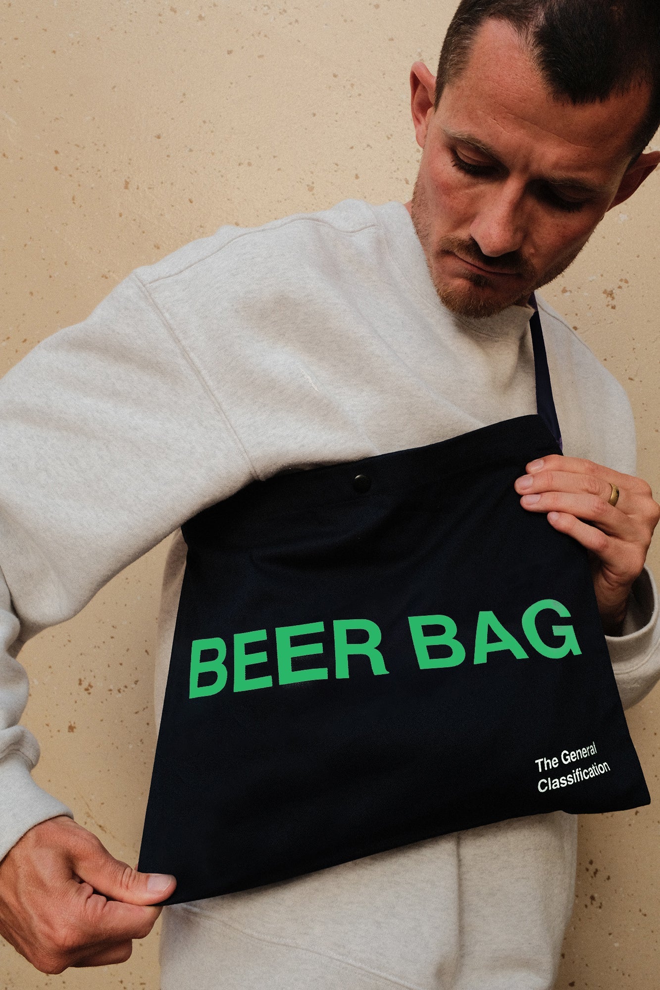 A man wearing a light grae sweatshirt holds up a black tote bag with bright green text reading 'BEER BAG' and small white text reading 'The Great Classification.' against a beige textured wall.