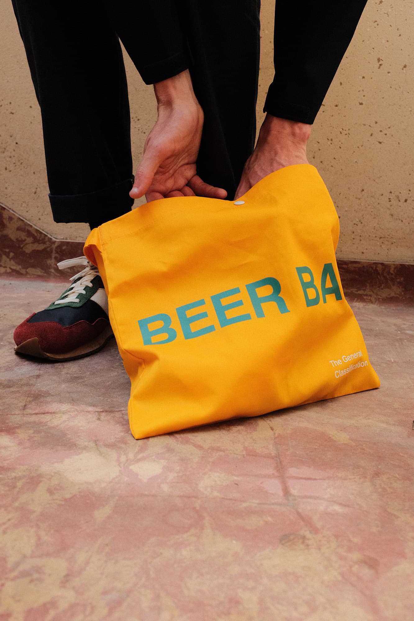 A person wearing black pants and a green-and-white sneaker holds a bright yellow canvas tote bag printed with the words 'BEER BAR' in blue. The scene is set on a textured terracotta floor against a beige wall.