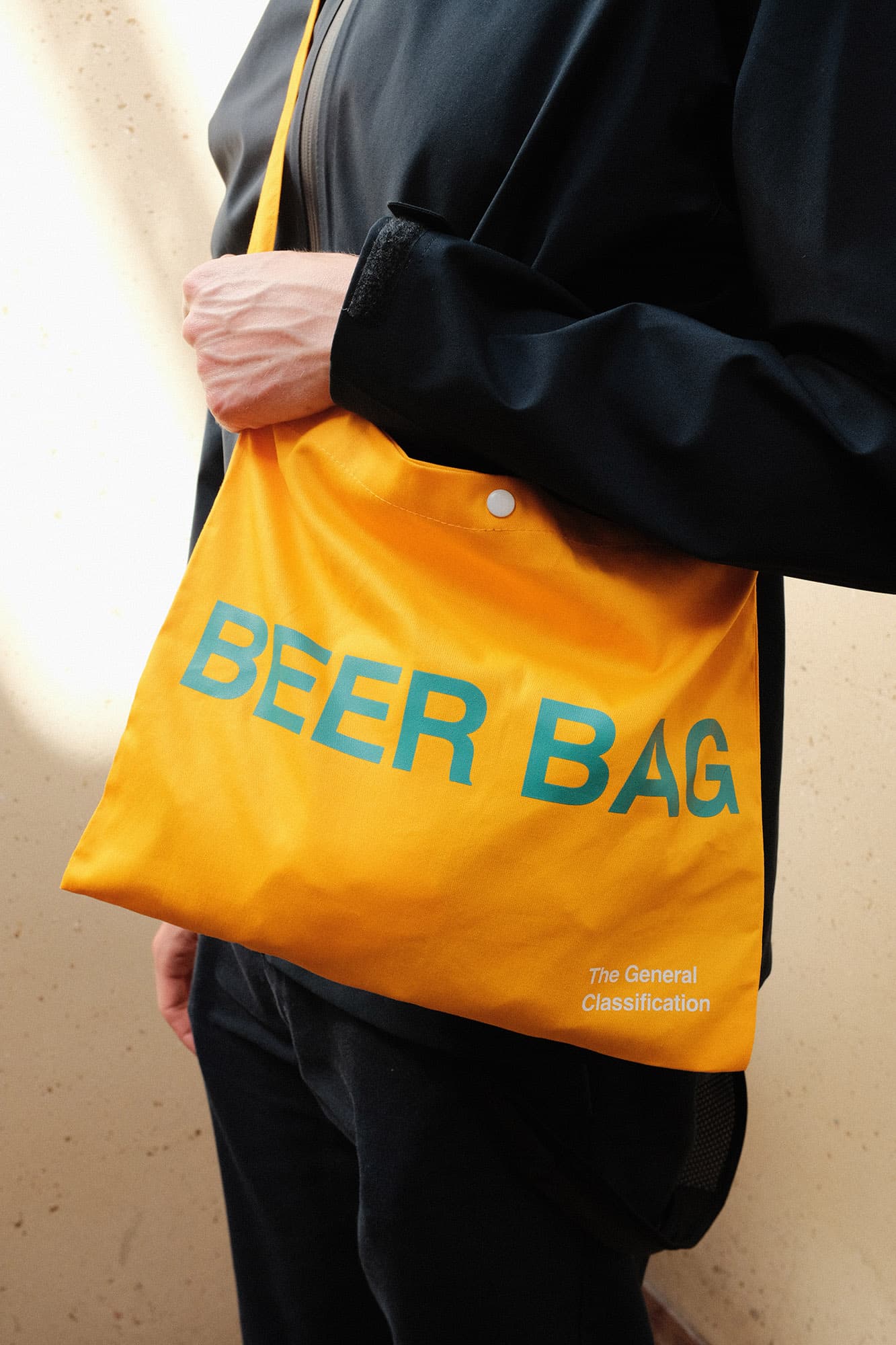 A person wearing a black jacket and black pants carries a bright yellow canvas shoulder bag with the words 'BEER BAG' printed in blue across the front. The bag also has small white text reading 'The General Classification' near the bottom right corner.