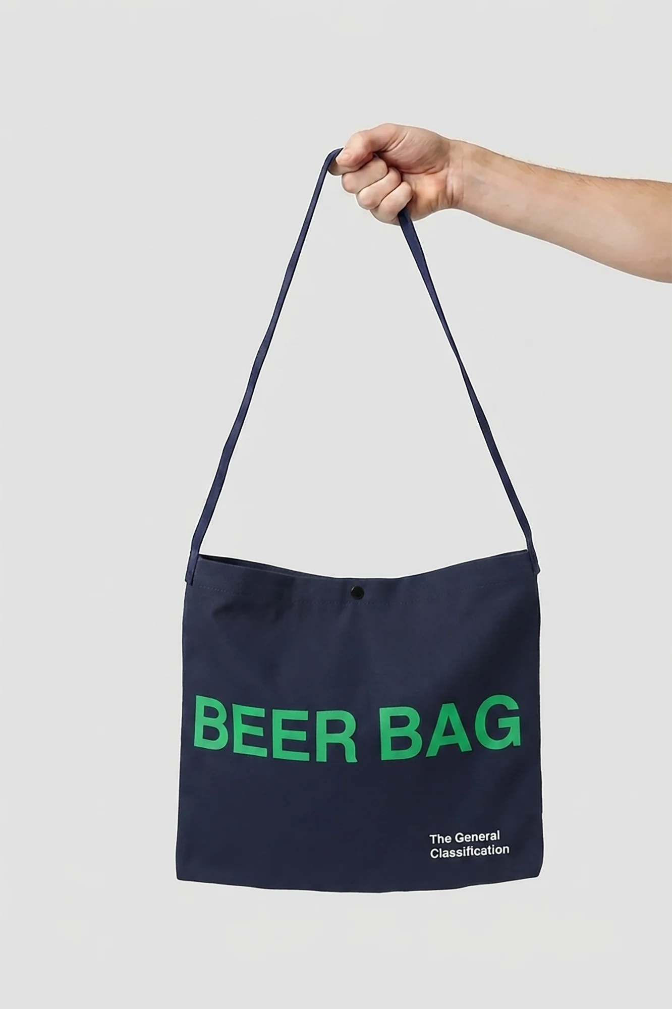 A navy blue canvas shoulder tote bag with green 'BEER BAG' text and small white 'The General Classification' text, held by a hand against a light gray background.