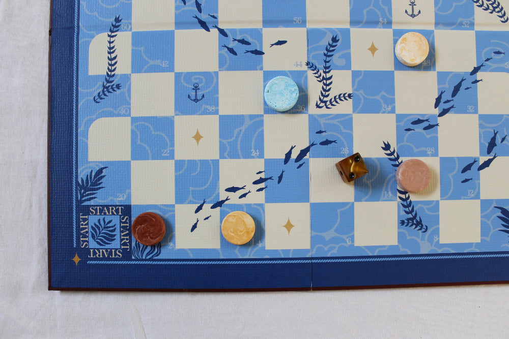 Close-up of the blue and cream Snakes and Ladders board featuring fish, anchors, seaweed, marbled counters and dice.
