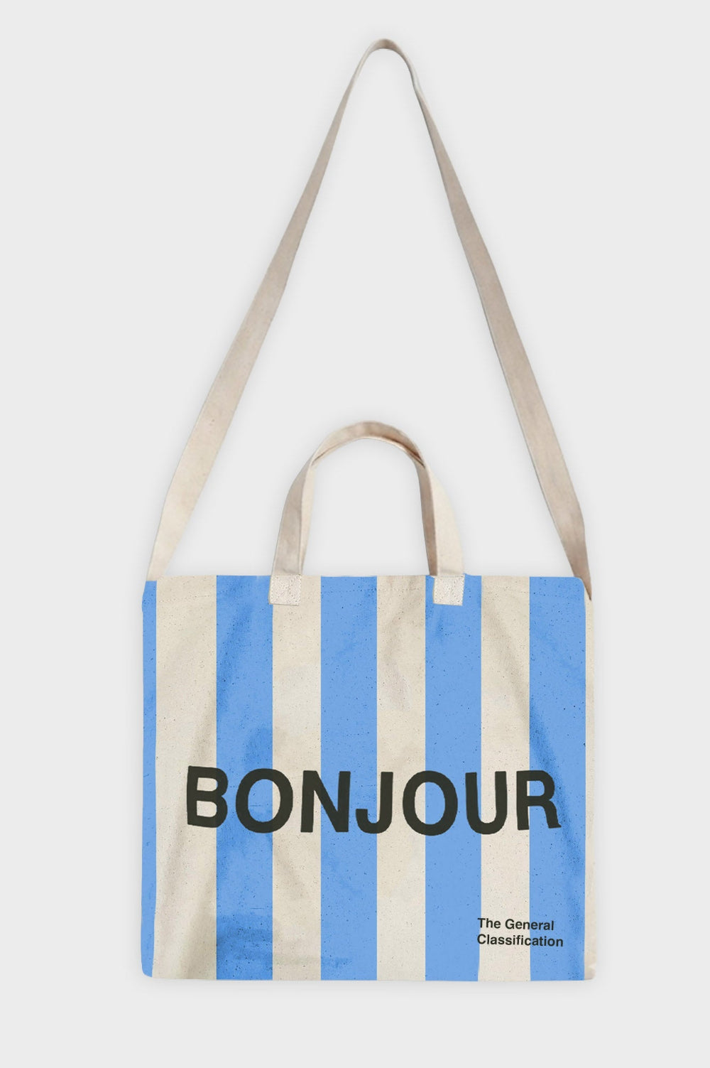 A canvas tote bag with vertical light blue and cream stripes, featuring the word 'BONJOUR' printed in bold black letters across the center and 'The General Classification' in small black text at the bottom right. It has two cream fabric handles and is photographed against a light gray background.