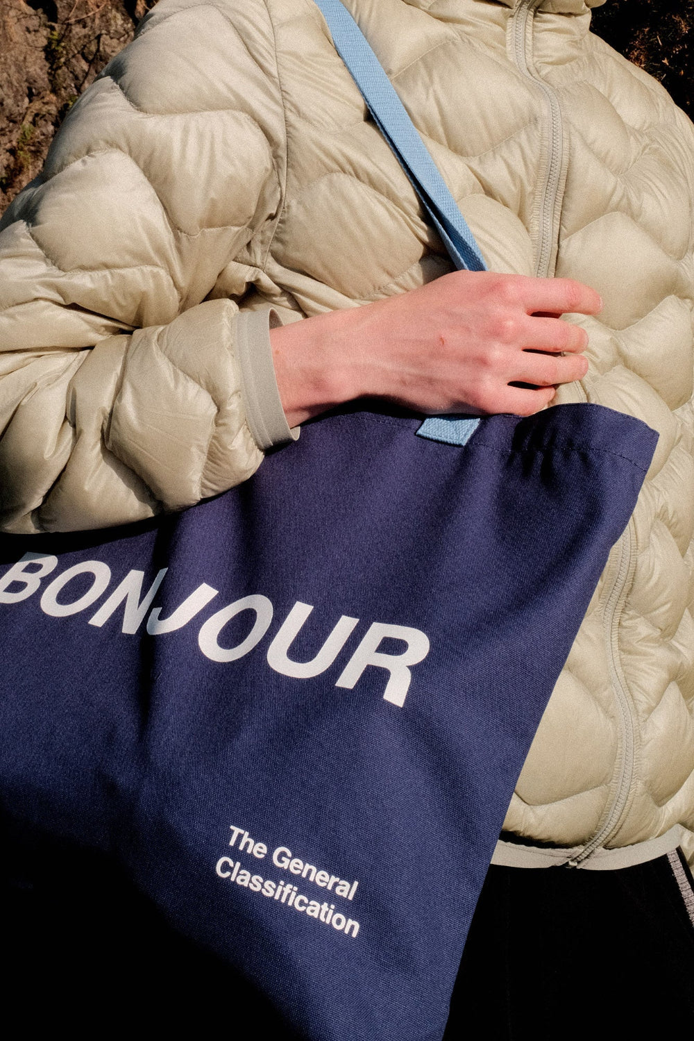 Person holding a navy tote bag with 'Bonjour' text, wearing a beige puffer jacket.