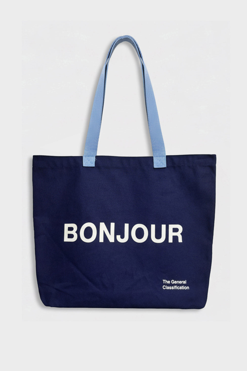 A navy blue canvas tote bag with light blue shoulder straps, featuring the word 'BONJOUR' printed in bold white letters on the front, with 'The General Classification' in smaller white text below, set against a plain light grey background.