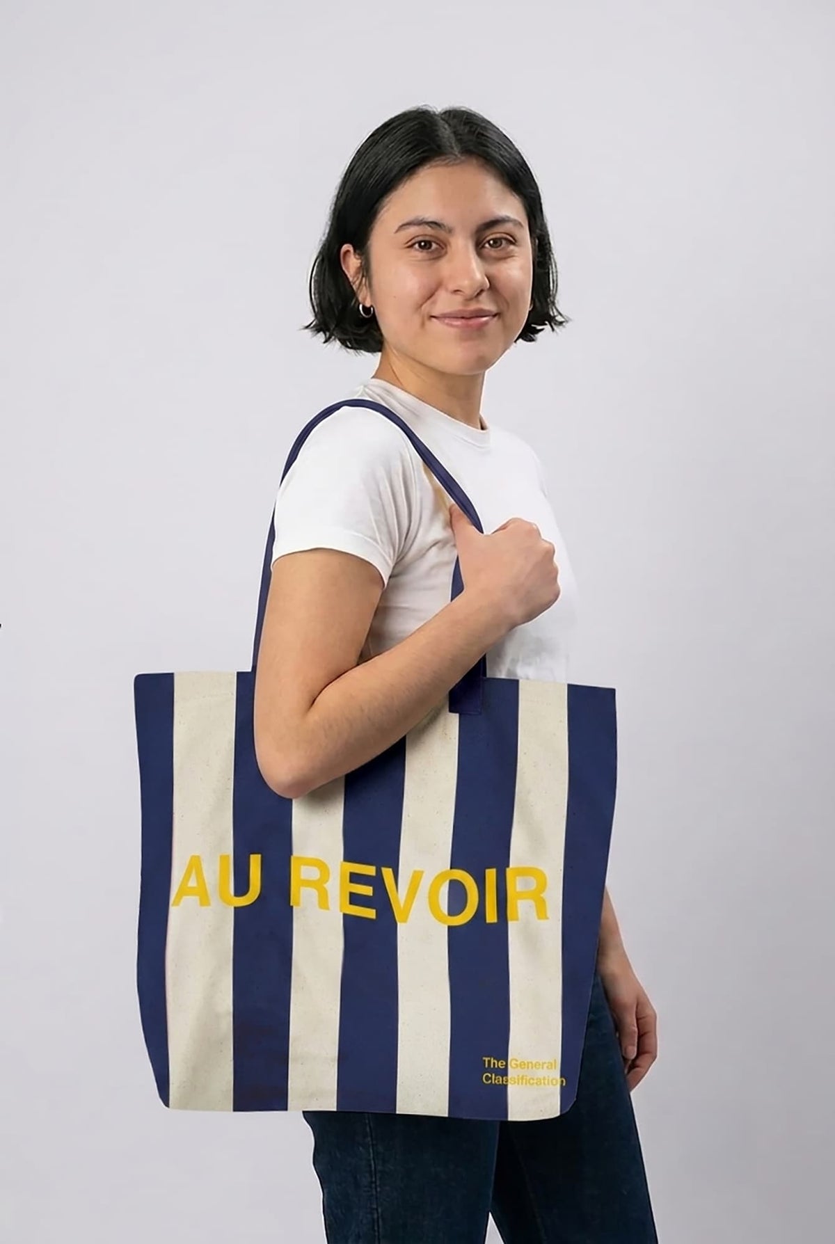 Woman holding a striped tote bag with 'AU REVOIR' text on a plain background