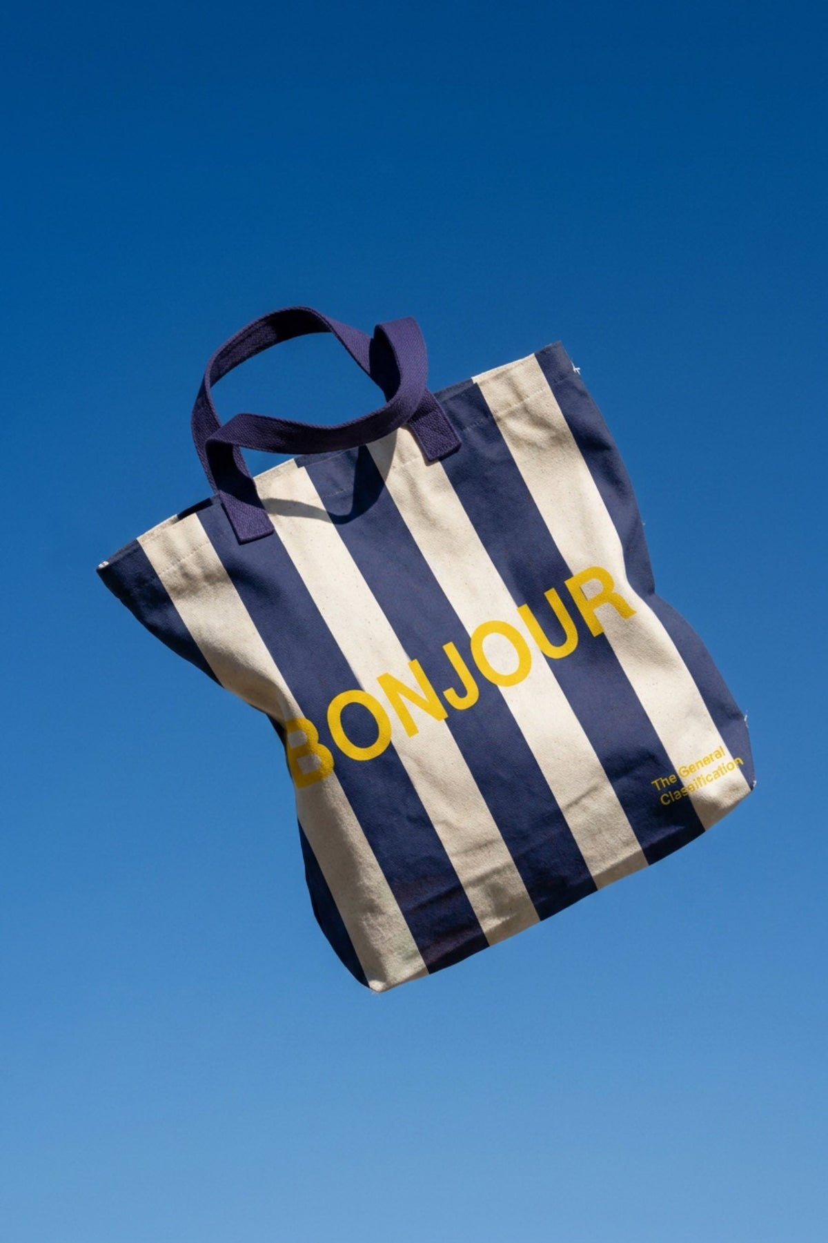 Navy and beige striped tote bag with 'Bonjour' text against a clear blue sky
